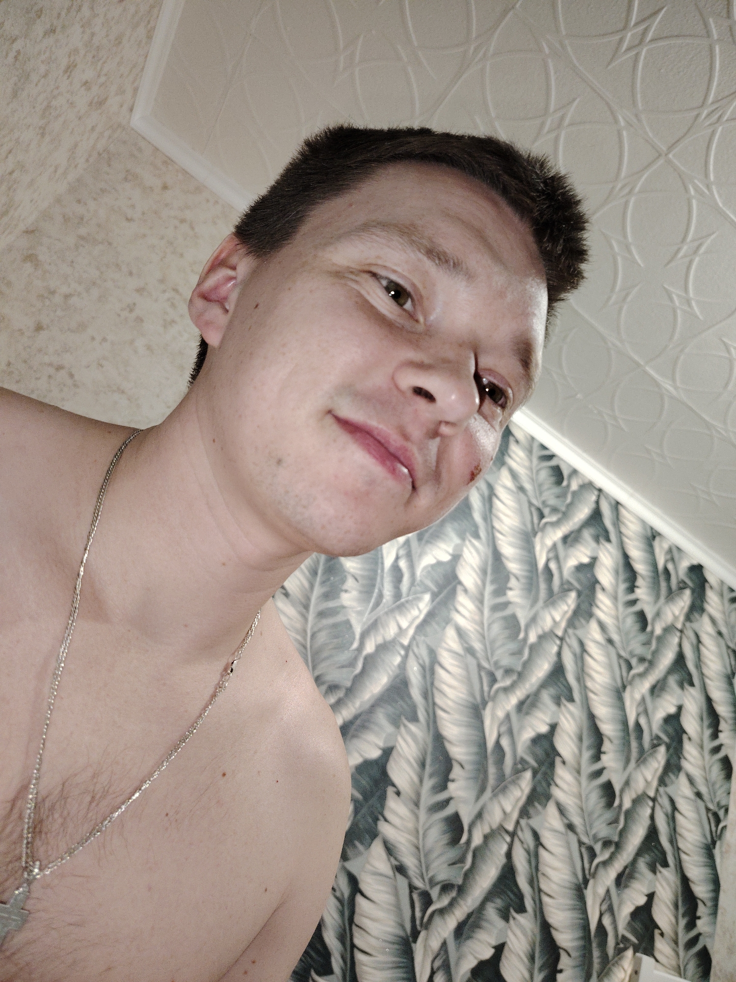 This image depicts an adult male subject in an interior setting, located in the city of Марковский, Russia. The subject is shown from a low-angle perspective, from the chest up, and is shirtless. He has short, dark hair and light skin with visible freckles. A small, dark red scab is present on the left side of his nose, near the nostril. A faint red mark is visible on his upper lip. Light facial stubble is apparent on his chin and cheeks. He is wearing a silver-colored chain necklace with a visible cross pendant on his chest, where sparse chest hair is also present. The subject's head is tilted slightly, and his eyes are directed towards the right side of the frame, with a subtle upward curve at the corners of his mouth. The background consists of three distinct surfaces: a light-colored, subtly textured wall to the upper left, an off-white ceiling with an embossed geometric pattern to the upper right, and a wall covered with grey and white stylized leaf-patterned wallpaper to the lower right. A white cornice separates the ceiling and the leaf-patterned wall.