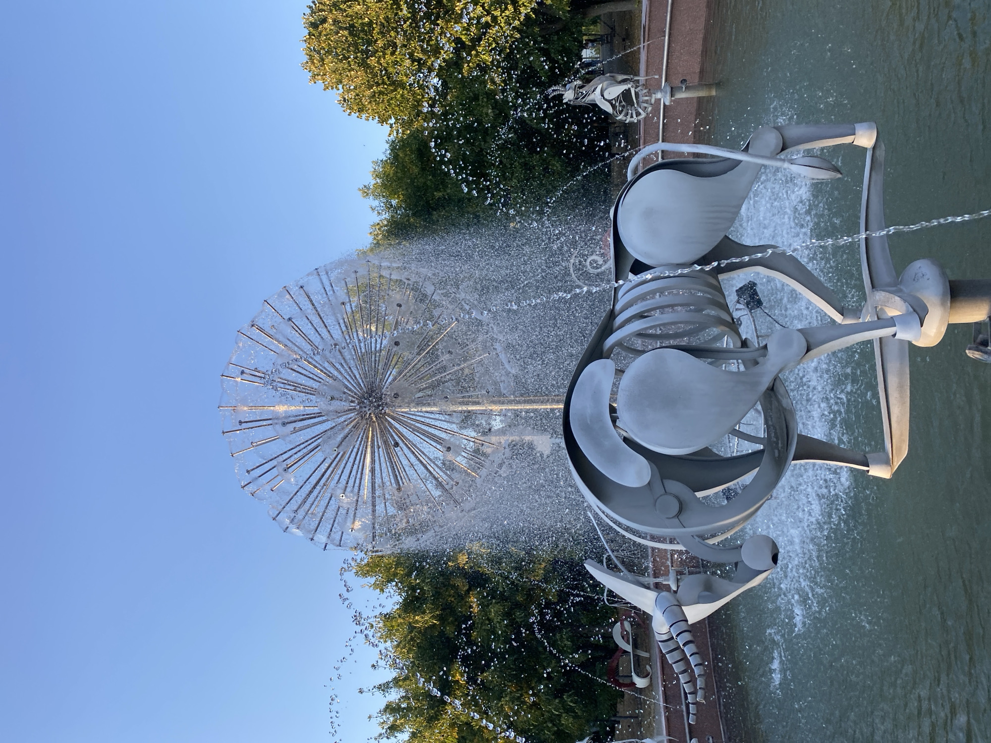 The photograph shows a large, modern fountain sculpture depicting a skeletal bull in the foreground. The bull is made of sleek, metallic material and is partially submerged in the water of the fountain. Behind the bull, a spherical, dandelion-like fountain sprays water upwards and outwards, creating a shimmering curtain. The background features lush green trees and a clear blue sky. In the middle ground, other similar metallic sculptures, possibly other skeletal animals, are visible. The scene is set in the city of Chișinău, Moldova, likely in a public park or square. The lighting suggests it is daytime, possibly late afternoon given the warm tones in the sky and on the foliage. There are no people visible in the immediate vicinity of the sculptures, but there are benches and paths that suggest this is a frequented public space.