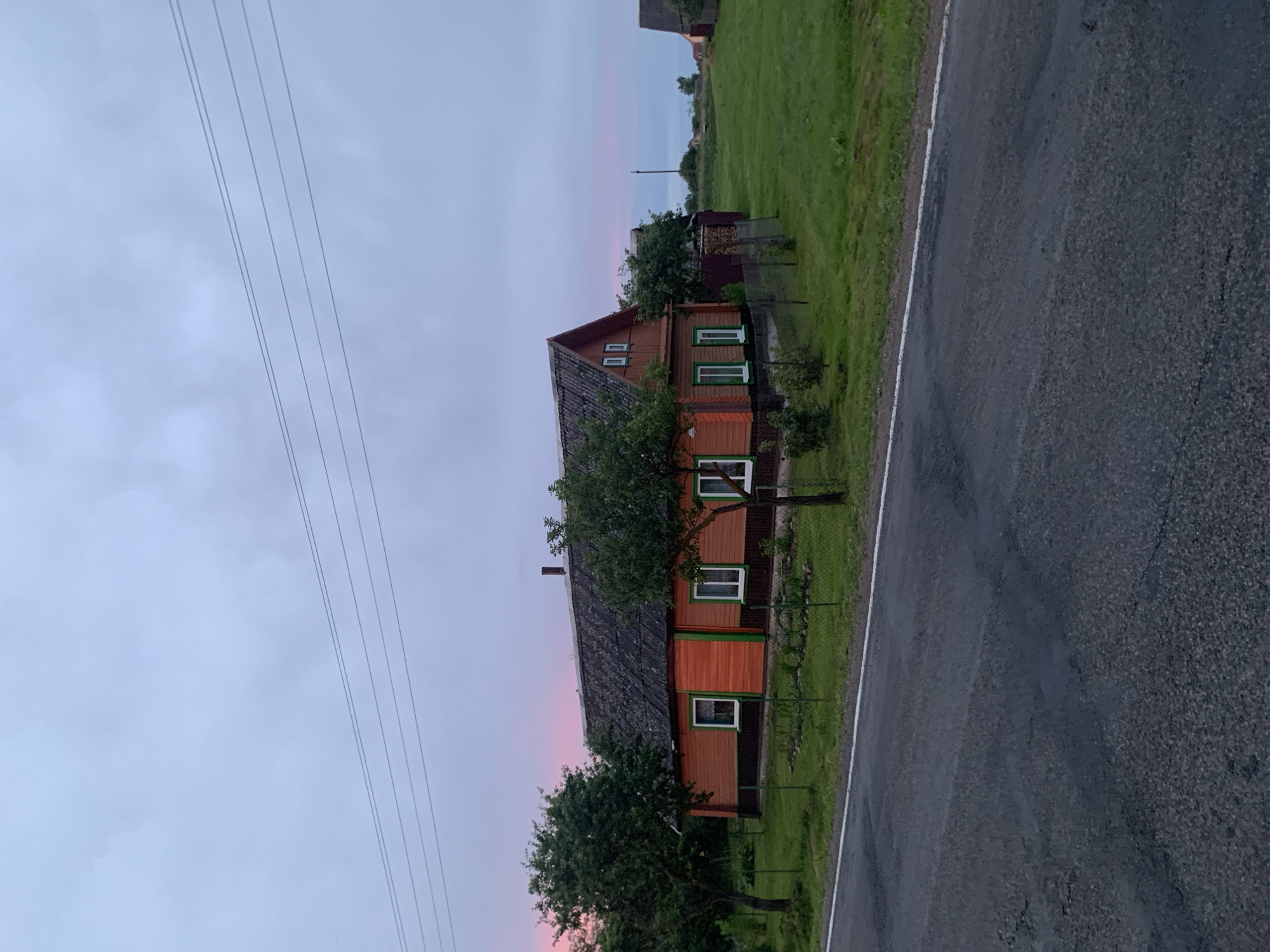 The image depicts a rural scene in Відзаўскі сельскі Савет, Belarus, at dusk. A two-story wooden house with an orange exterior and green trim is the central focus. The house has a gray, textured roof and several white-framed windows, including two square windows on the upper gable. A tall, dark chimney is visible on the roof. In front of the house, a small garden area with rocks and greenery is enclosed by a metal fence. Two mature trees with dense green foliage stand on either side of the house, and a few smaller saplings are planted in the garden. A wire fence runs along the edge of the property. The foreground shows a dark asphalt road with a white line marking its edge, suggesting it is a local road. The background consists of a vast, green lawn and more distant trees and buildings, with a hint of a pink and purple sunset sky visible in the upper left corner. Overhead, power lines stretch across the frame. The overall impression is one of quiet, rural evening in a Belarusian village.