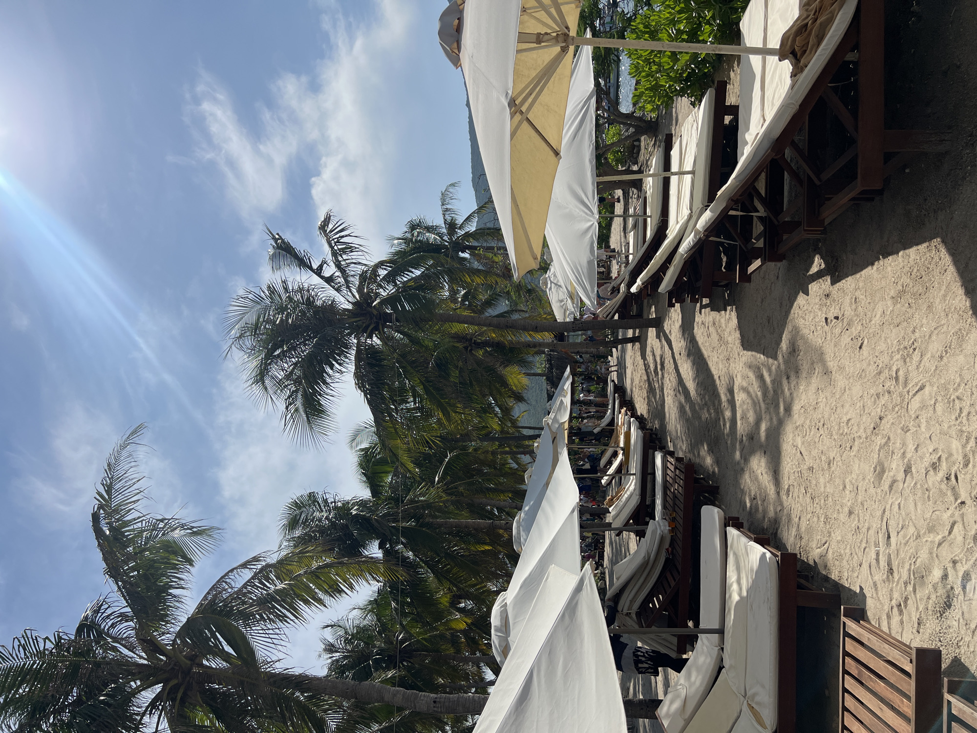The image shows a beach scene in Nha Trang Ward, Vietnam. The foreground contains a sandy beach lined with wooden lounge chairs covered in white cushions, each sheltered by a white or off-white umbrella. Palm trees are interspersed along the edge of the beach, their fronds reaching toward a blue sky with scattered clouds. A bright light source, possibly the sun, is visible in the upper left corner, creating a lens flare. In the background, hints of vegetation and possibly the sea can be seen.