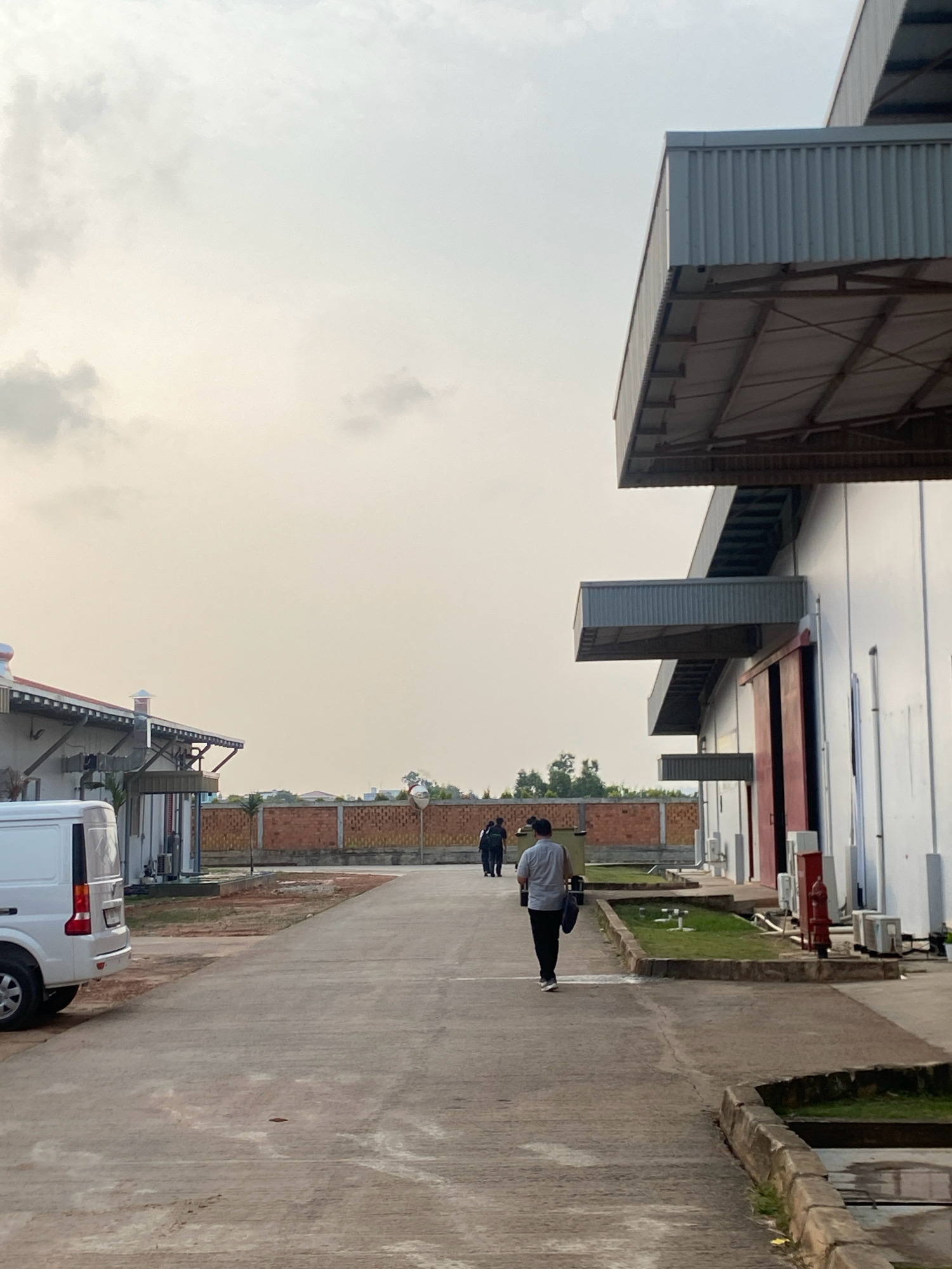 The image depicts a scene at the entrance of an industrial facility, likely in the city of Batam, Indonesia, judging by the architecture and vegetation. The sky is overcast with a pale, diffused light. In the foreground, a concrete pathway leads into the facility. To the left, a white van is parked. A man in a grey shirt and black pants is walking away from the camera down the pathway, carrying a dark bag. Further down the path, two other figures are visible.
The industrial buildings are characterized by white walls, corrugated metal roofs, and large red doors. Overhanging structures with corrugated metal siding are prominent features of the buildings. A brick wall with a chain-link fence runs along the right side of the pathway, separating the facility from the surroundings. Green grass and some small trees are visible in patches. To the right, fire hydrant and multiple outdoor air conditioning units are installed along the building wall. In the distance, more buildings and some trees are visible. The overall impression is one of an active but quiet industrial environment.