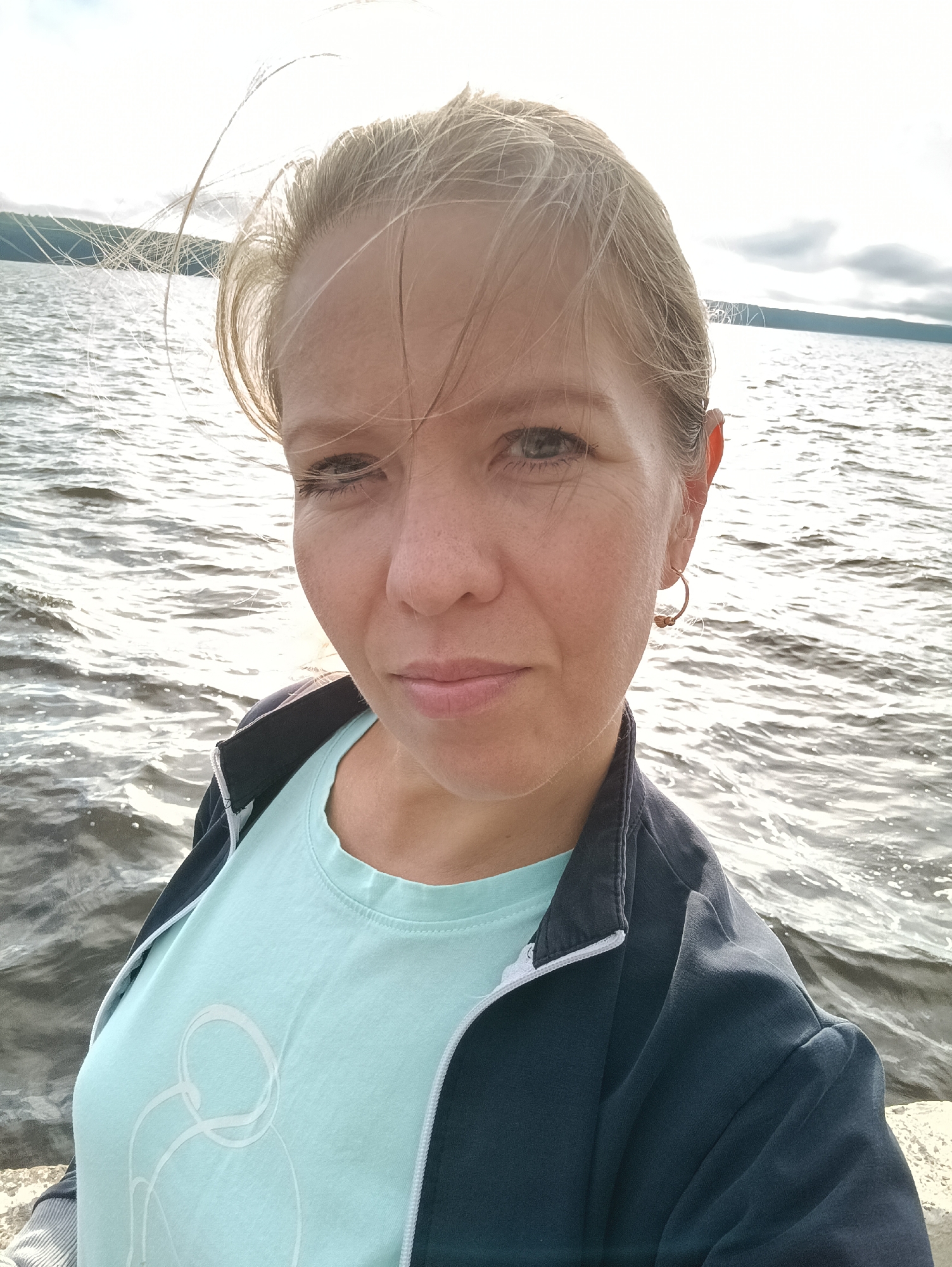 A medium shot photograph captures a woman with blonde hair posing for a selfie outdoors. She is wearing a light blue t-shirt with a white abstract design and a dark blue jacket with a white zipper. The woman has light skin, fair eyebrows, and blue-gray eyes. She is wearing a hoop earring in her left ear. Her hair is light blonde and appears to be blowing in the wind.
The background of the image is a body of water, likely a reservoir or large river, with choppy waves reflecting the sunlight. In the distance, a dark, tree-lined shore is visible under a cloudy sky with patches of bright sunlight. The overall lighting suggests it is daytime.
The location is identified as being near the Kama River in Chaikovsky, Russia. The woman is standing on what appears to be a concrete structure or embankment, possibly a pier or a retaining wall, by the water's edge. The scene conveys a sense of being outdoors in a natural, possibly recreational, setting near a body of water.