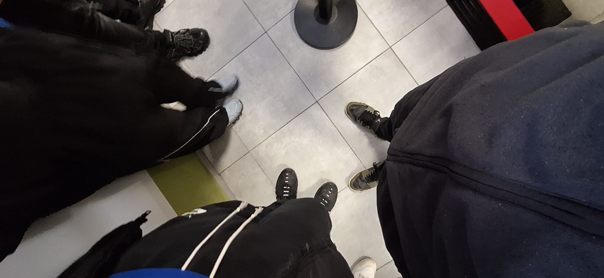 The image shows a ground-level view of three people standing on a tiled floor. The leftmost person is wearing dark clothing with a white stripe down the leg, and grey and black shoes. The person in the middle is wearing black shoes with white stripes and black pants with white drawstrings. The person on the right is wearing dark clothing and dark sneakers. A black circular object is visible in the background. The setting appears to be indoors. There is insufficient information to verify a location in Burgas, Bulgaria.