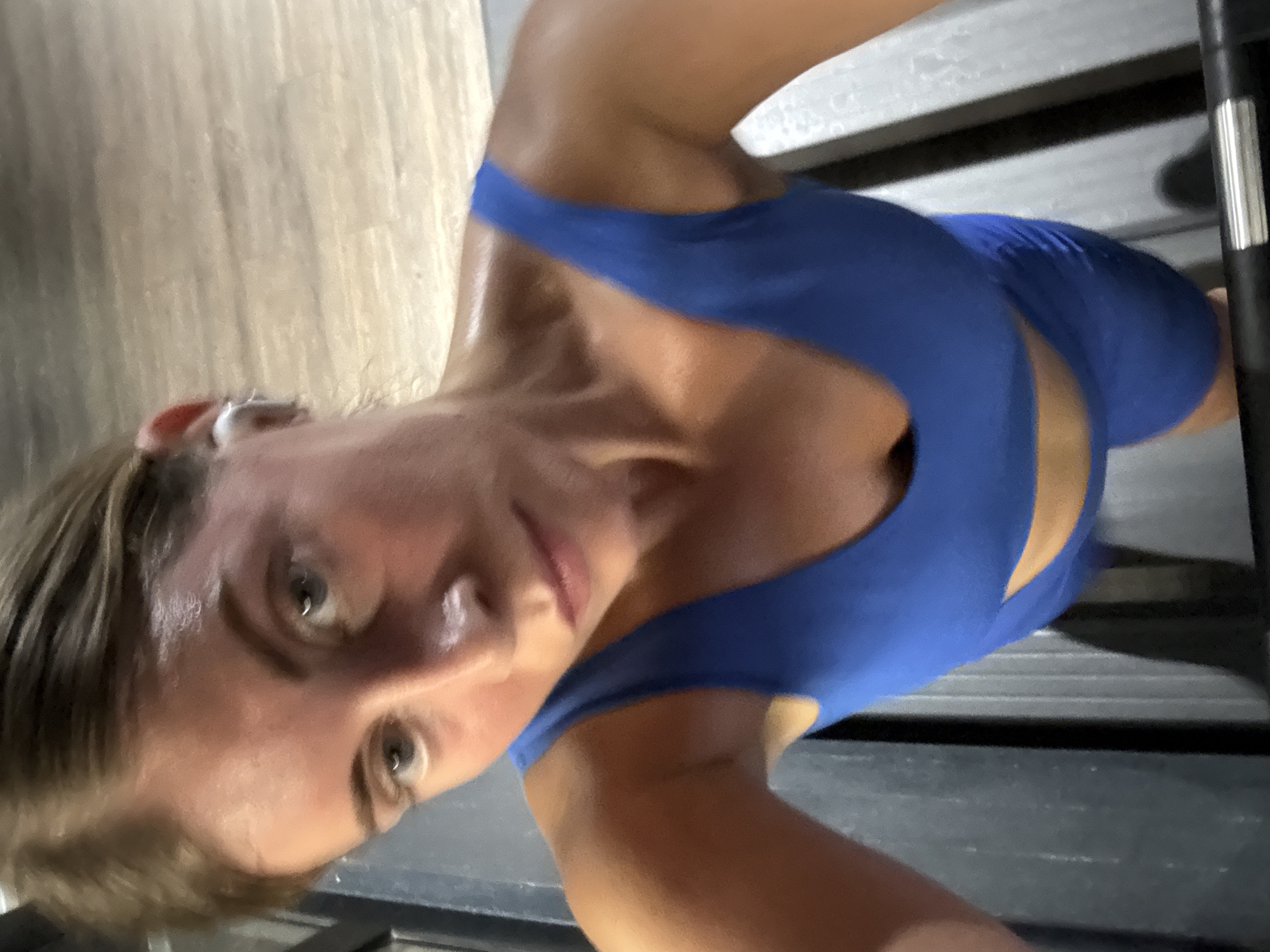A woman is captured in a close-up, high-angle selfie as she exercises on a treadmill. She is wearing a blue athletic top and appears to be sweating, indicating a strenuous workout. Her expression is focused and determined. The background shows the textured surface of the treadmill belt and a wooden floor, suggesting an indoor gym setting. The lighting is dim, with a warm, slightly yellow cast. There is no visible text in the image. The scene implies a personal fitness activity in a gym.