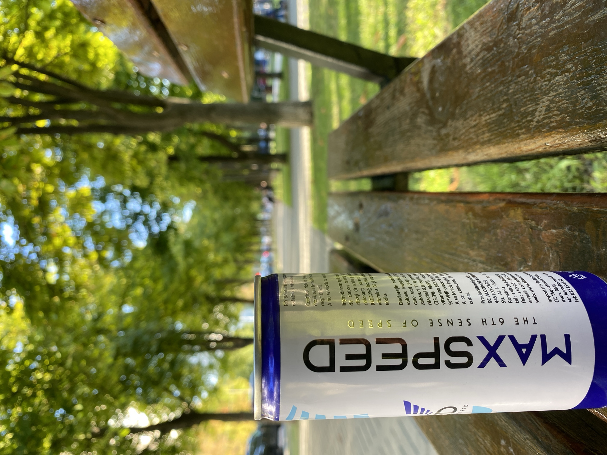 This is a close-up, eye-level, outdoor shot of a can of "MAXSPEED" energy drink, placed on a weathered wooden park bench. The can, predominantly white with blue accents and the prominent "MAXSPEED" logo in large purple letters, is positioned horizontally. The text on the can is oriented vertically and is in Romanian, with nutritional information and ingredient lists visible.
The background is softly blurred, suggesting a park or a tree-lined area. Green foliage and the dappled sunlight filtering through the leaves indicate it's daytime, likely late morning or afternoon, with pleasant weather. The shallow depth of field focuses attention on the energy drink, making it the clear subject of the image. The overall mood is casual and relaxed, consistent with a park setting. Visible text on the can includes "MAXSPEED THE 6TH SENSE OF SPEED," and various Romanian phrases related to ingredients and nutritional values, alongside manufacturer and importer details for Moldova.