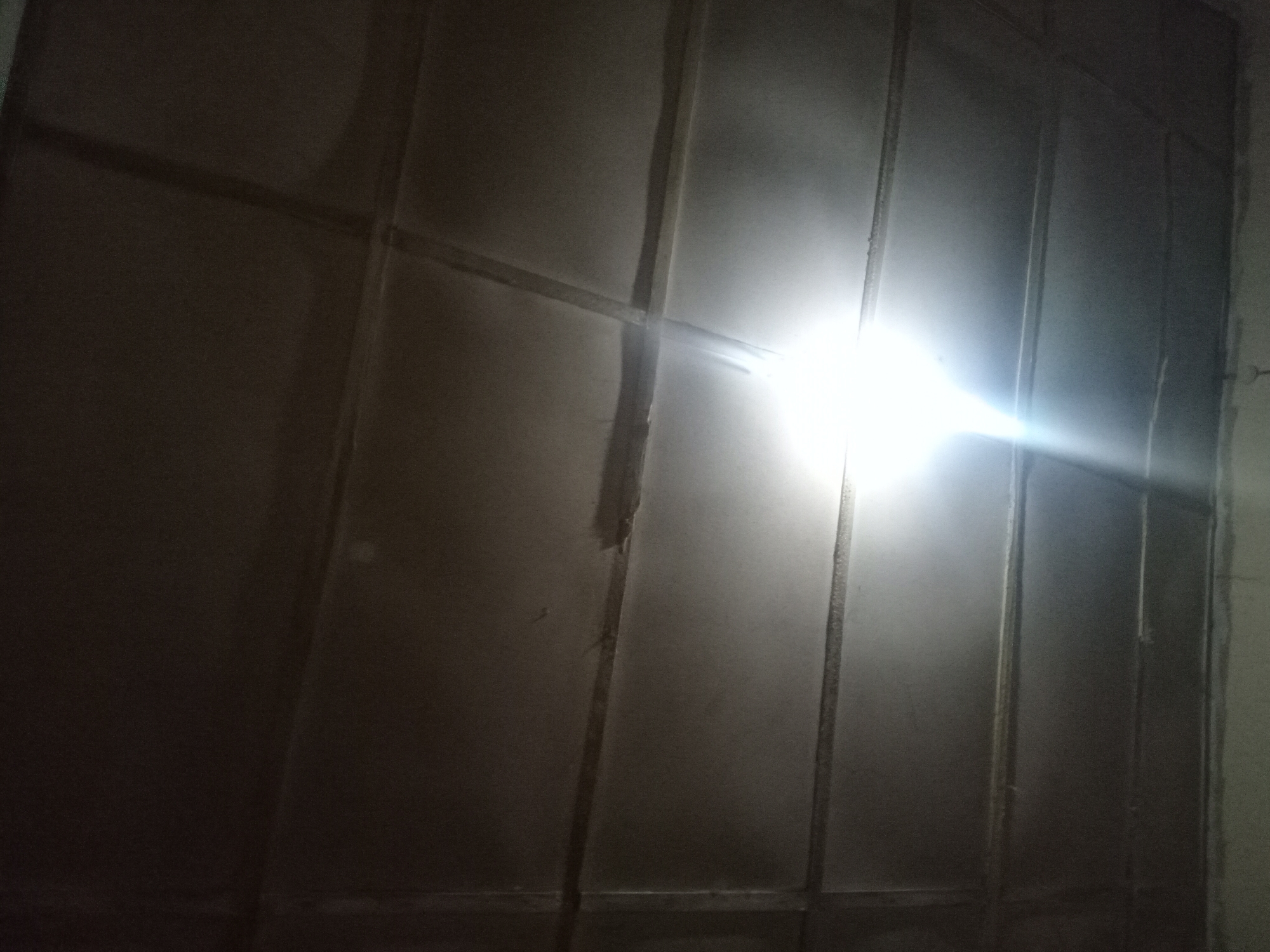 The image shows a low-angle view of a dark, textured ceiling with horizontal and vertical beams creating a grid pattern. A bright, white light source, possibly a light fixture, is positioned centrally, casting a strong glare and illuminating sections of the ceiling. The overall impression is of an indoor space with artificial lighting. There are no people or other discernible objects in the frame. The environment appears to be dark, with the light source being the primary source of illumination. The ceiling itself seems to be constructed of panels.
