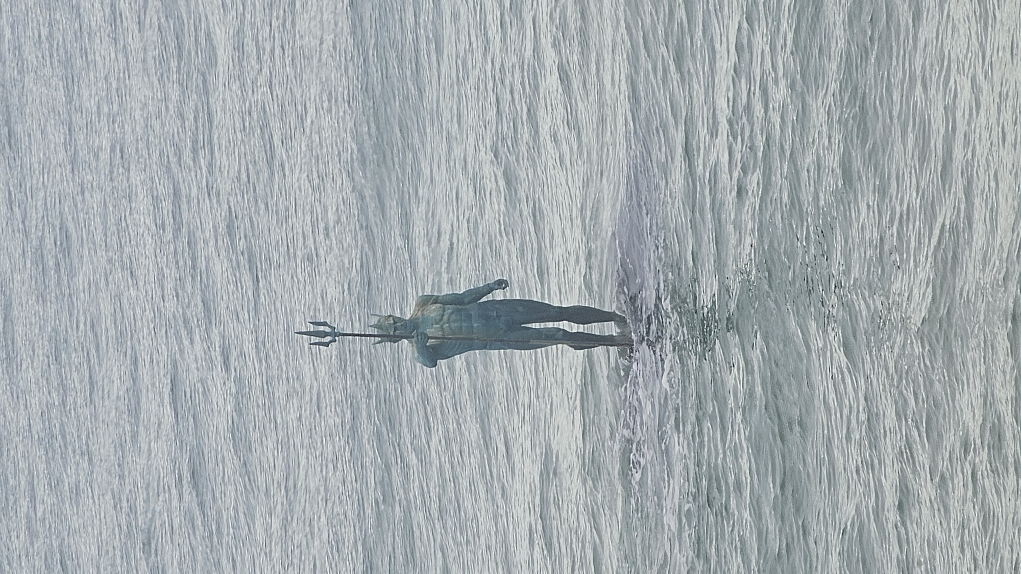 A statue of Neptune is displayed against a water wall backdrop. The statue is in the center of the image. The scene takes place in Varna, Bulgaria.