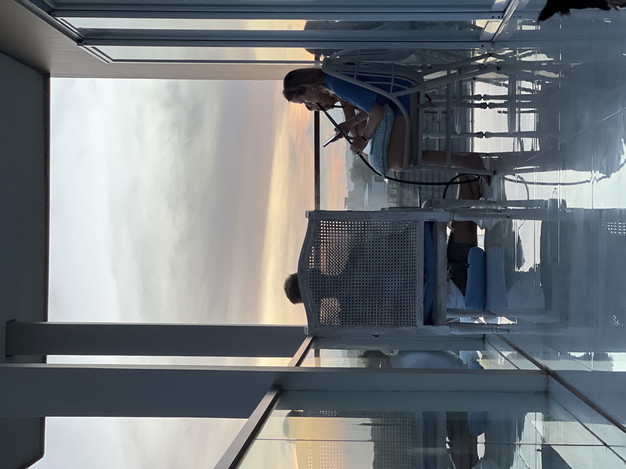 Two people are seated on white patio chairs on a high-rise balcony overlooking a city at sunset. The foreground showcases architectural elements of the building, including white columns and glass panels, suggesting a modern design. One person is facing toward the city and appears to be using a cell phone, while the other person is seated behind and slightly to the side, their features partially obscured by the chair's design. The city is shrouded in a golden hue from the sunset. The balcony appears to have a glossy, reflective floor, adding to the image's modern aesthetic.