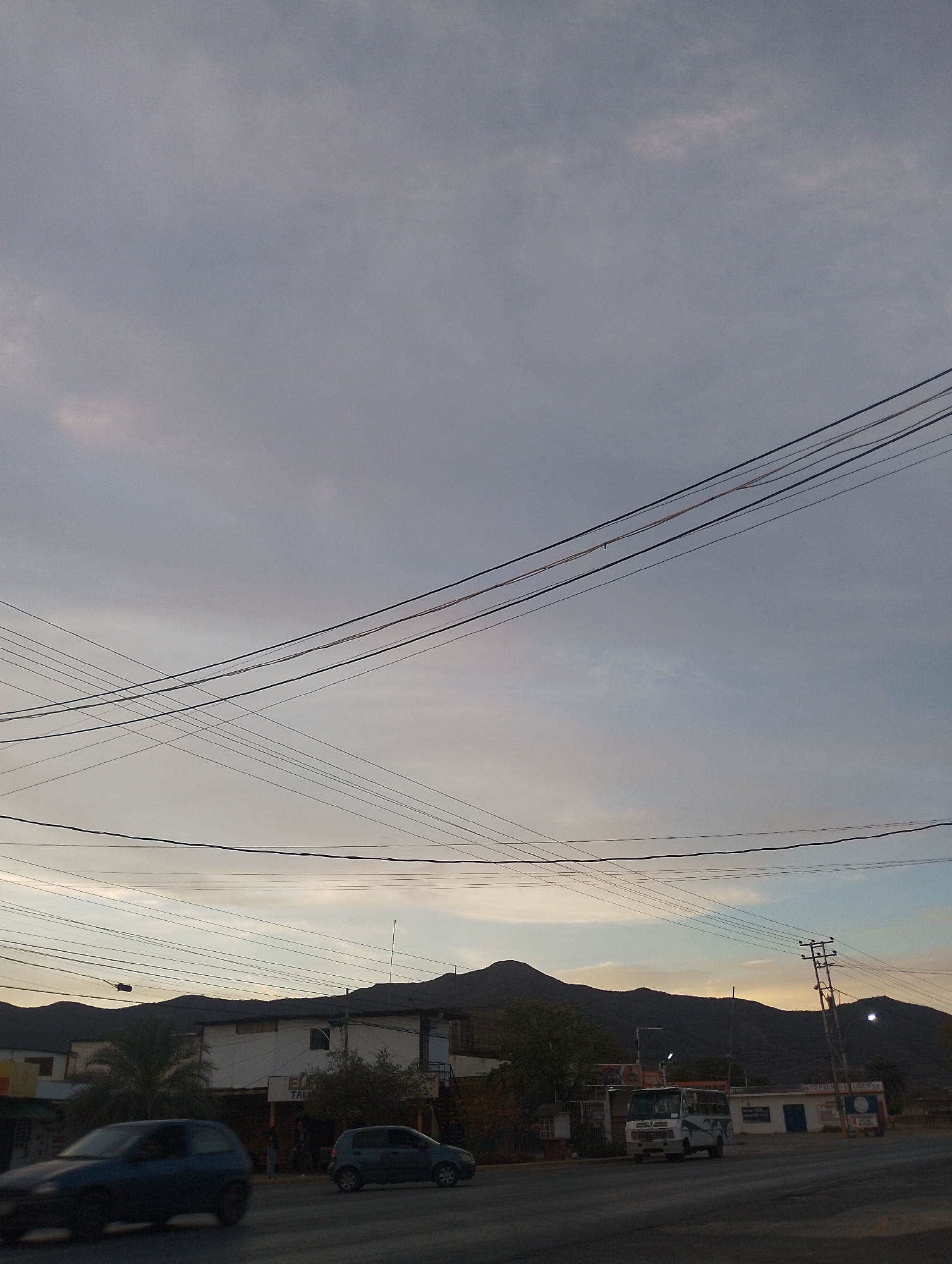 The image captures a street scene in Las Barrancas, Venezuela, featuring a cloudy sky with power lines crisscrossing the frame. In the midground, there are buildings, a palm tree, and parked cars. A mountain range is visible in the background. A light-colored vehicle is on the street on the right. The focus is on the buildings and natural landscape, with the sky and power lines providing context.