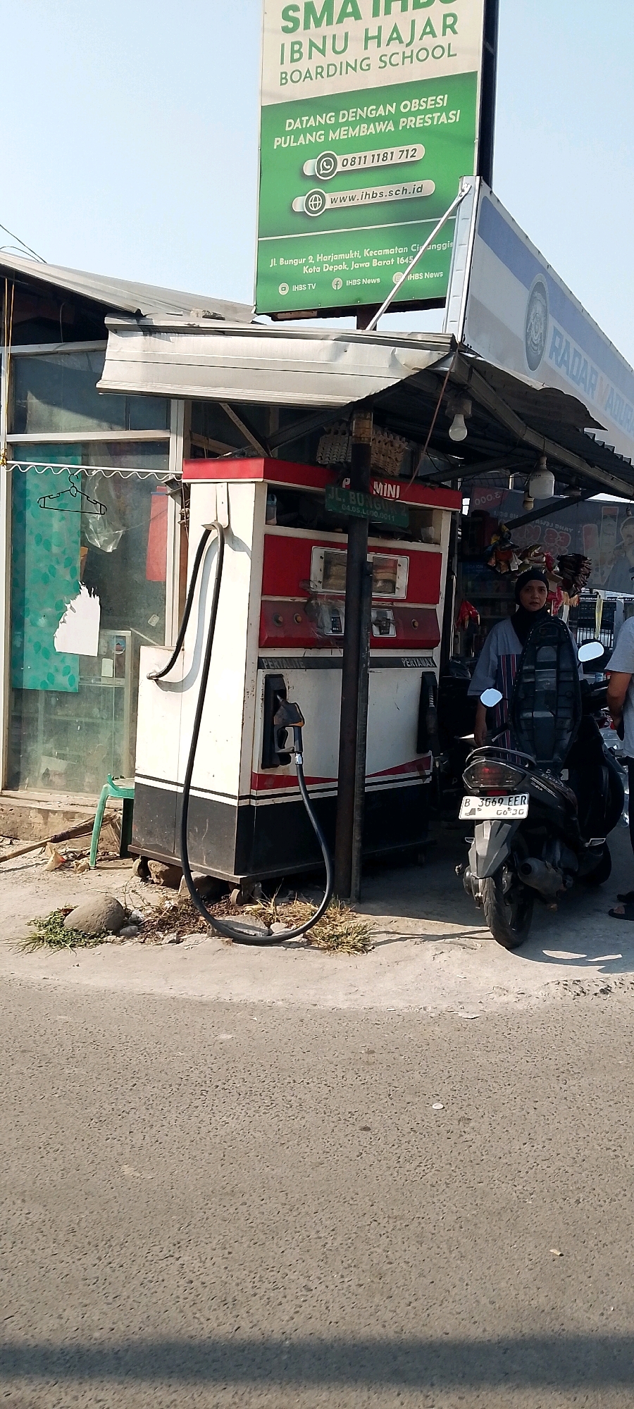 The image depicts a street scene in Depok, Indonesia, likely on a sunny day, given the harsh shadows. The focal point is a small roadside gas station, characterized by a red and white fuel pump with a black hose. To the left of the pump, a glass-fronted shop or kiosk is visible, with some items displayed inside, including what appears to be a hanger on a rack. A green plastic stool is placed near the shop. A black motorcycle with a license plate reading "B 3669 EER" is parked in front of the gas station, and a person wearing a blue shirt and a headscarf is standing next to it, looking towards the camera. In the background, a large green billboard advertises "SMA IHBS IBNU HAJAR BOARDING SCHOOL" in Depok, providing contact information and a website address. The surrounding environment consists of a paved road, a sidewalk, and some vegetation and debris around the base of the gas station. Another smaller sign on a pole near the gas station reads "MINI JL. BUNGUR 2". The overall impression is a typical street view of a small business in an Indonesian urban setting.