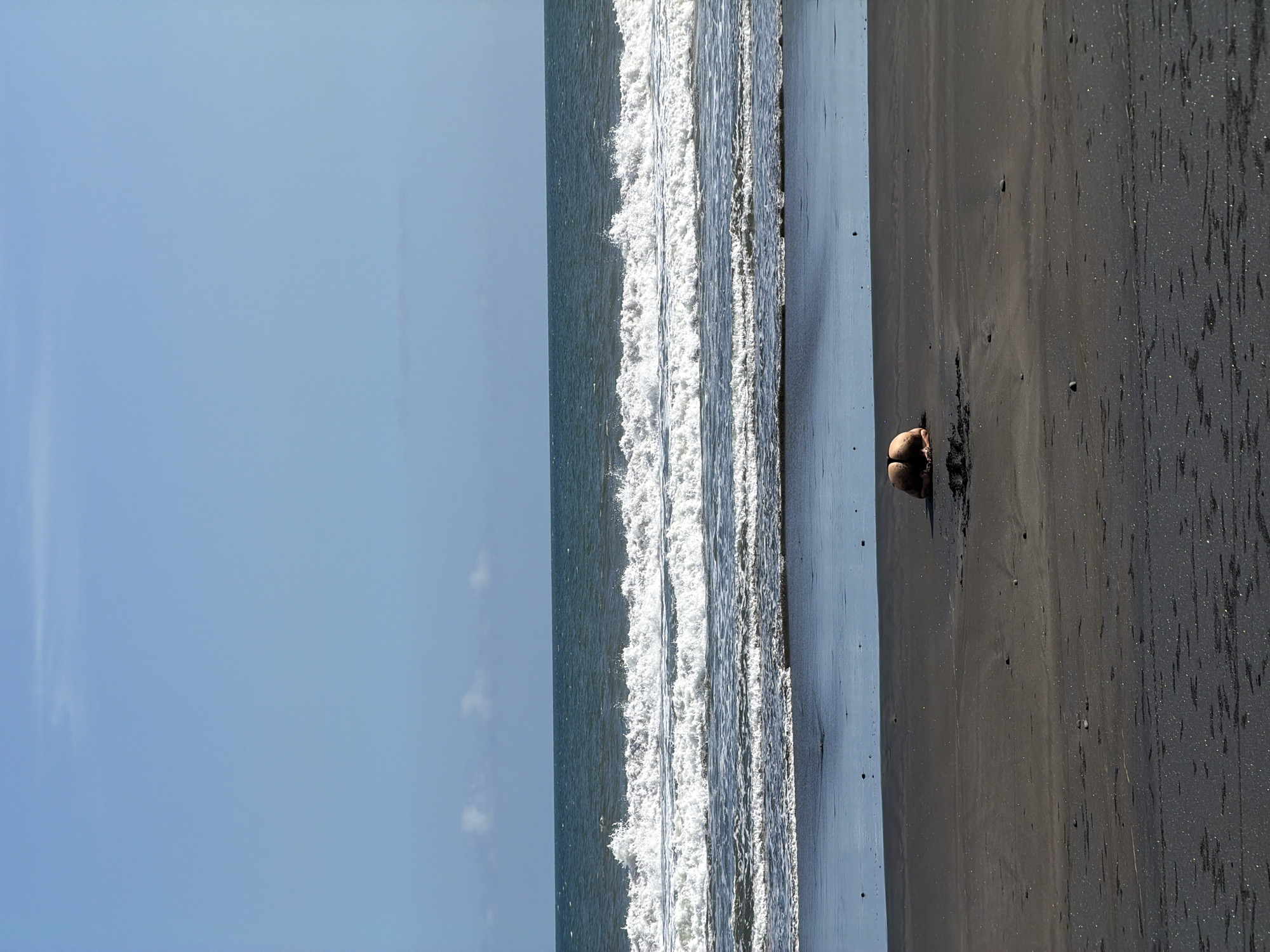 The image depicts a beach in Selemadeg Timur, Indonesia. A person is on the dark sand. The ocean's waves are visible. The sky is blue with a few clouds.