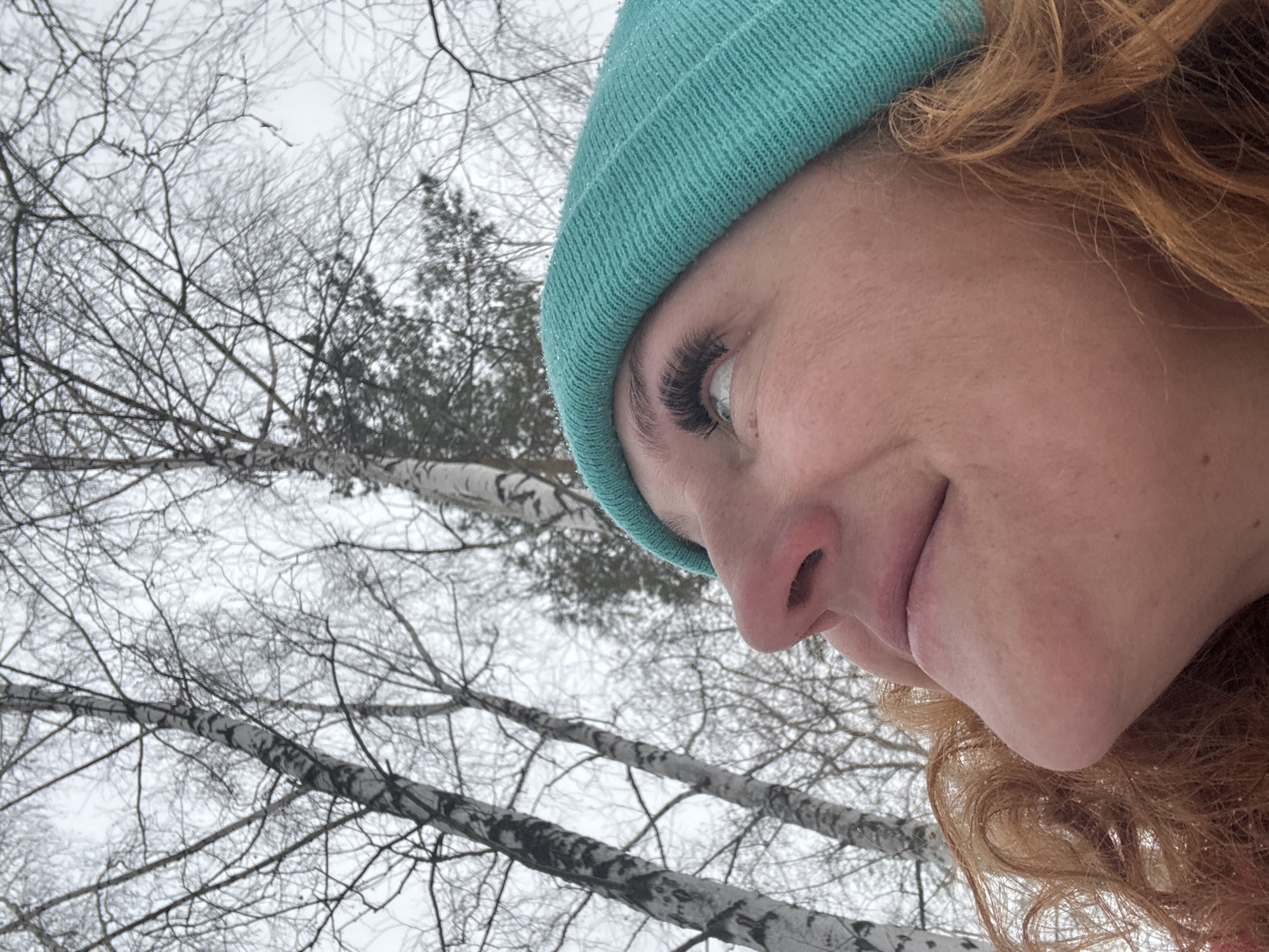 A close-up, upward-tilted photograph shows a person with fair skin, auburn curly hair, and a teal knitted beanie. The person's face is angled towards the camera. Enhanced eyelashes are visible on the person's left eye. The background features a wintry scene with bare trees, some of which appear to be birch trees with white bark. The sky is overcast and white, suggesting a snowy or cold day.
The city of Noviy, Russia is not discernible within the provided image.