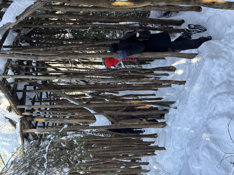The image displays an outdoor winter scene within a distinctive wooden structure in Kol'tsovo, Russia. The structure consists of numerous vertical, slender, debarked logs forming open walls and supporting a log-framed roof that is heavily covered in snow. The ground throughout the area is blanketed in deep, white snow, with a clearly defined pathway visible, worn through the snow and winding between the wooden columns. Bright natural light illuminates the scene, indicative of a clear winter day. Three individuals are present, walking along the cleared snow path, moving generally from right to left. The person in the foreground, on the right, wears a dark winter coat, dark pants, black winter boots, and grey and blue striped gloves. A dark backpack is worn, with a white cylindrical object attached to its side. A red knitted hat, featuring white eye-like accents, is visible on their head. A second individual, partially obscured in the mid-ground, wears a long, dark-colored coat and a plaid scarf. The third person, further in the background and largely obscured by the wooden columns, is also dressed in dark attire. All individuals are actively traversing the snowy pathway within the wooden installation.
