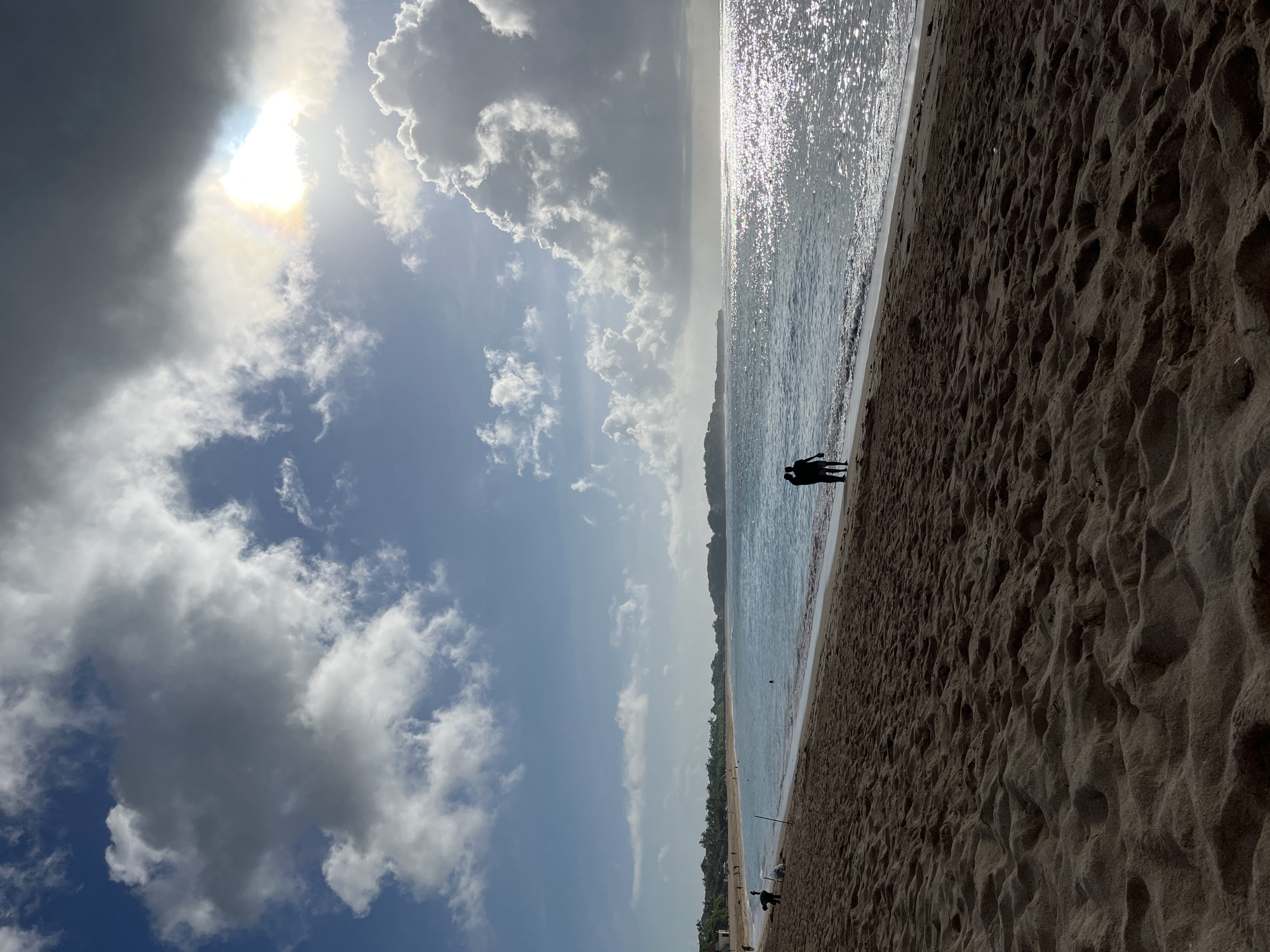 The image depicts a beach scene in Kuta Selatan, Indonesia, on a sunny day with a partly cloudy sky. The sun is shining brightly, partially obscured by clouds. The ocean is visible, with sunlight reflecting off the water surface. Two individuals are silhouetted on the shoreline, seemingly standing close to each other. The beach sand appears dark in color. There are also some trees and buildings further in the distance.