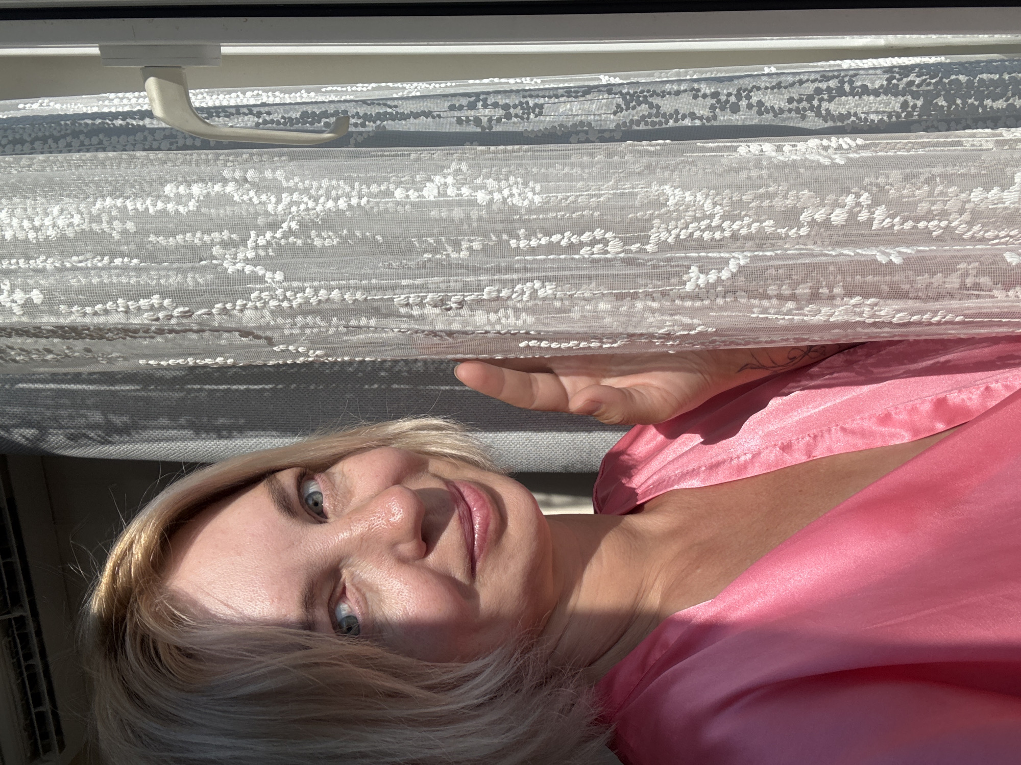 A woman with blonde hair and blue eyes, wearing a pink robe, stands by a window in Sochi, Russia. She is smiling and looking towards the camera. The window is covered by a sheer white lace curtain. Sunlight streams through the window, casting shadows on the wall and the woman's face. The woman's hand is visible, touching the curtain. The image appears to be taken indoors, with the woman positioned in the foreground and the window and curtain in the background.