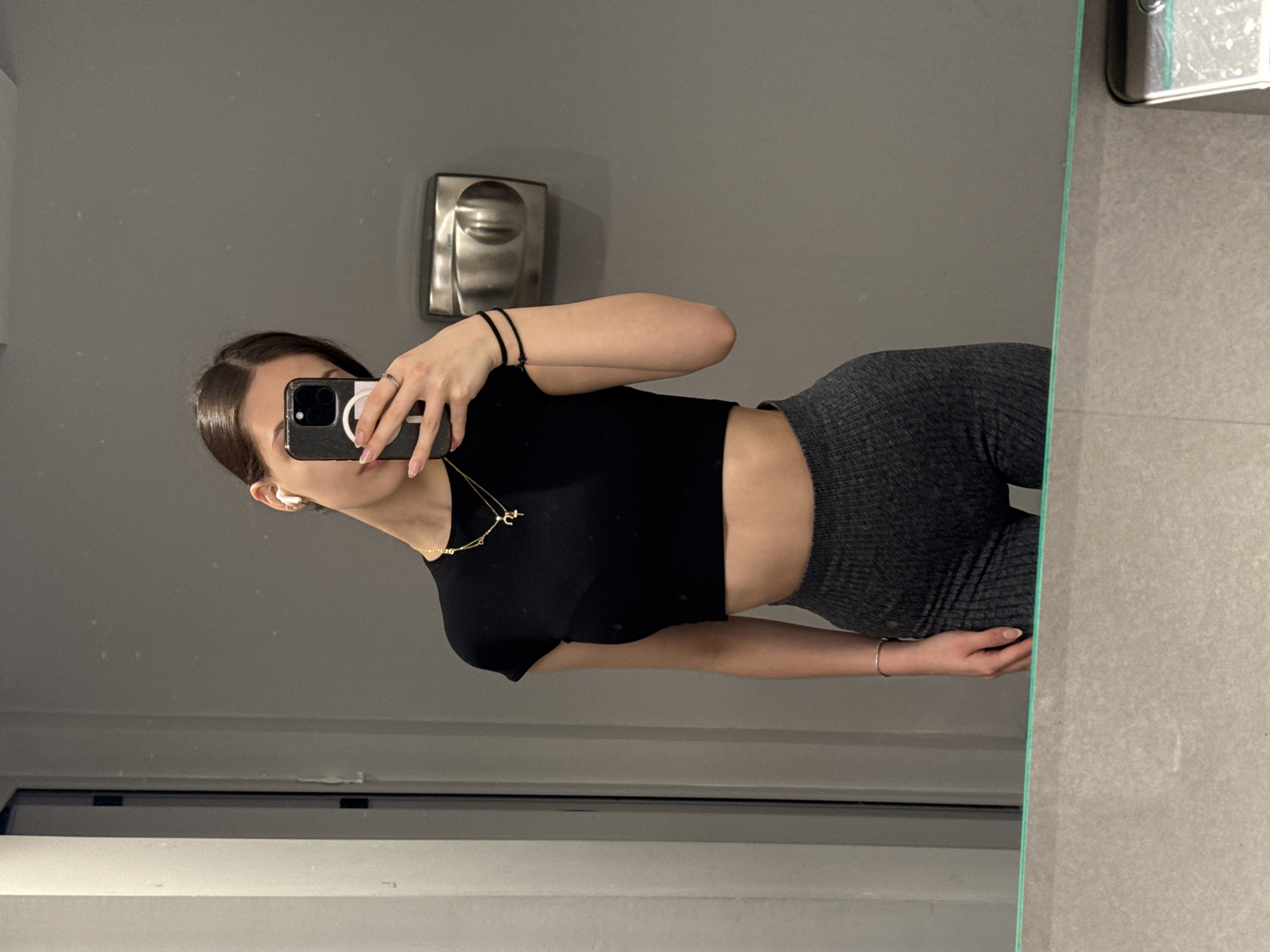 A young woman is taking a mirror selfie. She is wearing a black cropped top and dark gray leggings. A gold necklace is visible around her neck, and she has black hair pulled back. An object resembling a paper towel dispenser is visible behind her in the mirror. The background suggests a bathroom or similar facility. The location cannot be determined from the image without additional information.