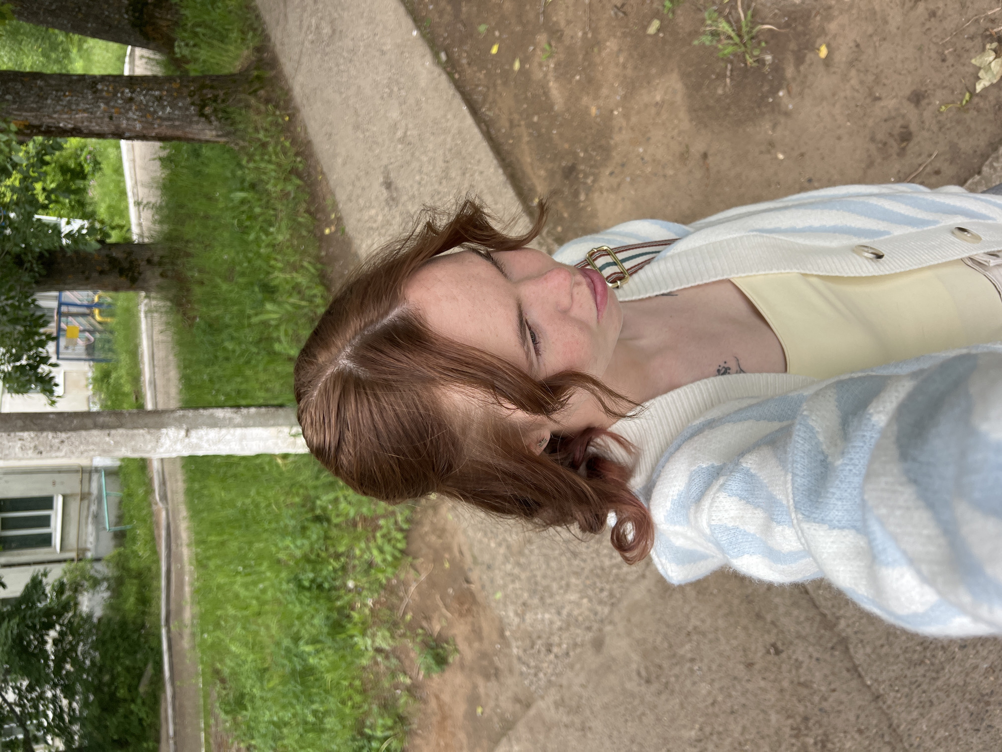 The image, rotated 90 degrees clockwise, captures a selfie of a young woman outdoors in Chaikovsky, Russia. She has reddish-brown hair with some waves and freckles visible on her face. Her eyes appear closed or downcast in the current orientation, but if the image were viewed upright, she would be looking upwards towards the camera. She is wearing a light yellow top under a cozy-looking white cardigan with light blue horizontal stripes. Buttons are visible on the front of the cardigan. A multi-colored striped strap (green, red, white) from a bag or garment is visible over her right shoulder. A small, dark tattoo of an intricate design is visible on her left forearm, just peeking out from under her cardigan sleeve.
The background suggests an urban or suburban outdoor setting. A concrete path or sidewalk runs through the scene, flanked by green grass and mature trees with textured bark. In the distance, parts of buildings or structures are discernible, including elements that suggest a playground with blue and yellow fixtures. The ground around the path is a mix of dirt and grass. The lighting indicates it is daytime, possibly with soft or indirect sunlight. There is no visible text in the image.
