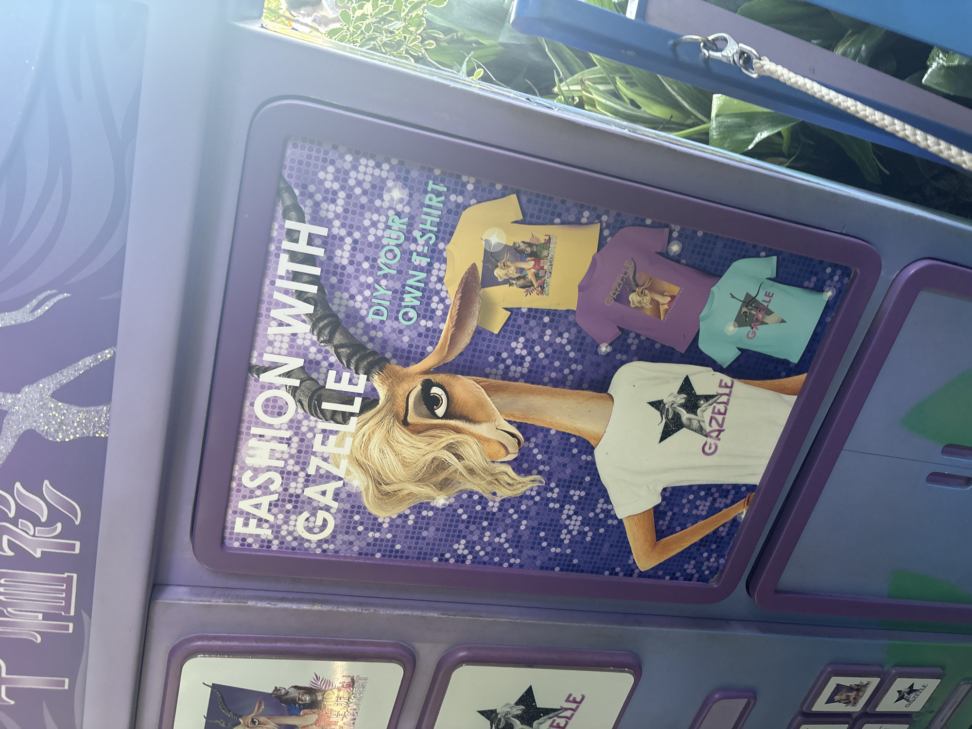 The image shows a purple display cabinet with various items related to a character named Gazelle. The cabinet is decorated with purple and white polka dots and features a large image of Gazelle. There are several T-shirts displayed, each with a different design featuring the character. The text on the display reads "FASHION WITH GAZELLE" and "DIY YOUR OWN T-SHIRT." Some of the T-shirts have images of Gazelle and the word "GAZELLE" printed on them. The cabinet is situated outdoors, with greenery and a rope visible in the background. The location is identified as Pudong, China.