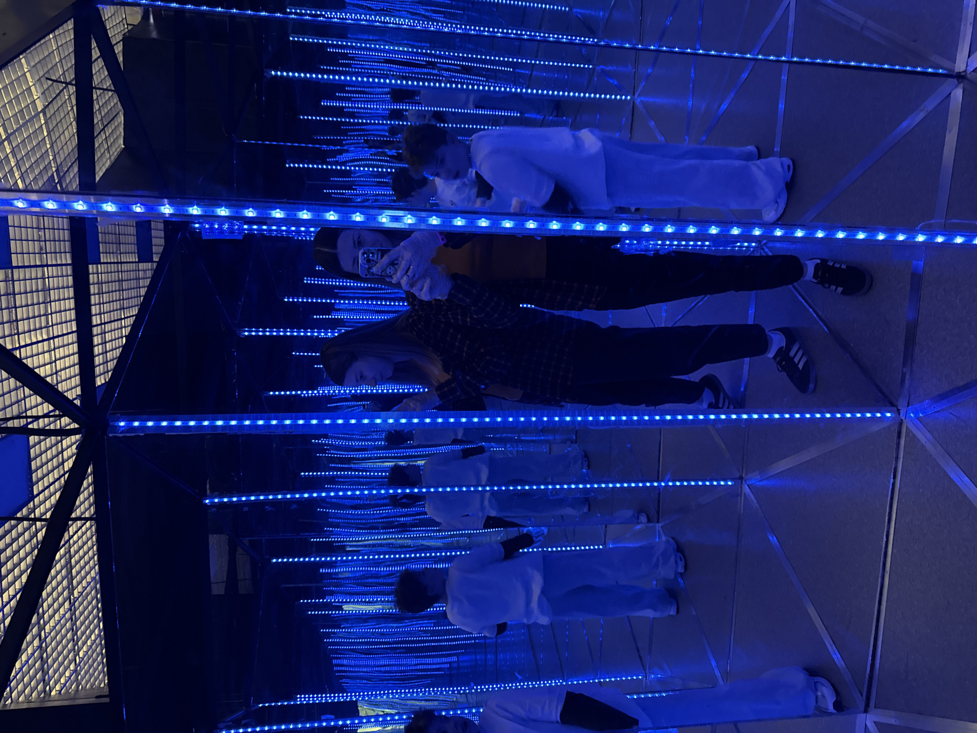 Two people are in a mirrored room with blue LED strip lights. The room appears to be part of an immersive art installation. One person is holding a phone and taking a picture of themselves and the other person. The location is Moscow, Russia.
