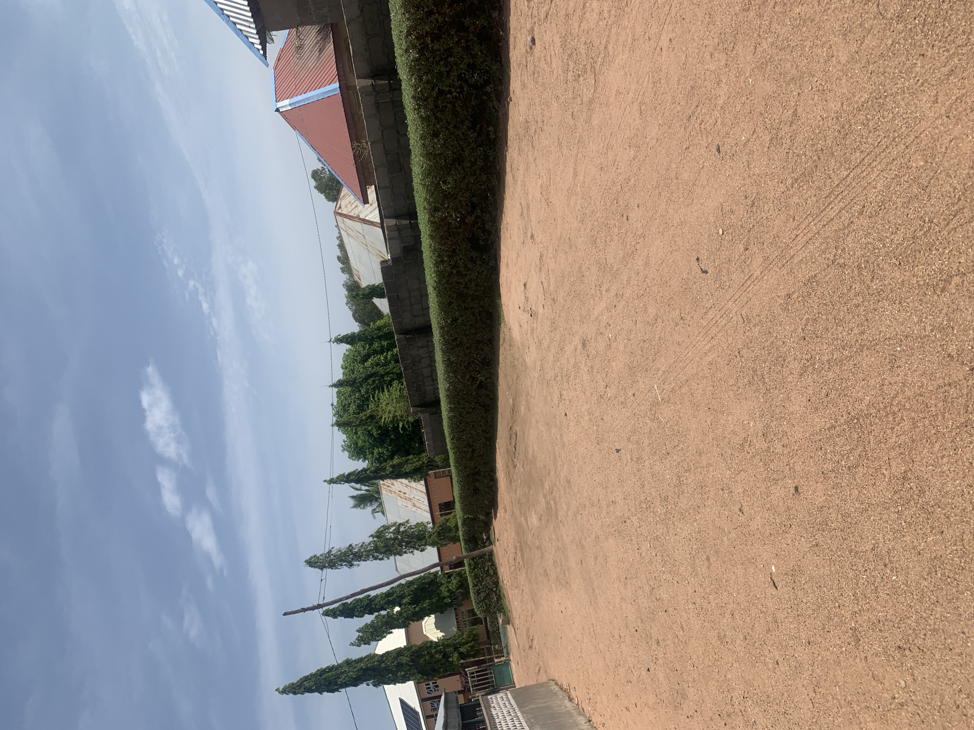 This image shows a street in Jauro Yinu, Nigeria. The street is unpaved and covered in reddish-brown dirt. On the right side of the image, there is a low, neatly trimmed green hedge that runs along a grey cinder block wall. Beyond the hedge and wall, there are several buildings with corrugated metal roofs, some painted red and blue, and others with a weathered, silver appearance. Tall, slender trees with dark green foliage are visible behind the buildings. On the left side of the image, a white textured wall is visible, with a green gate and fencing. Further back, a building with a white roof, possibly with solar panels, and the facade of another building painted in a light orange or peach color are partially visible. Power lines stretch across the upper portion of the image, supported by poles. The sky is blue with scattered white clouds. The overall scene depicts a residential or community area in a tropical or subtropical climate.