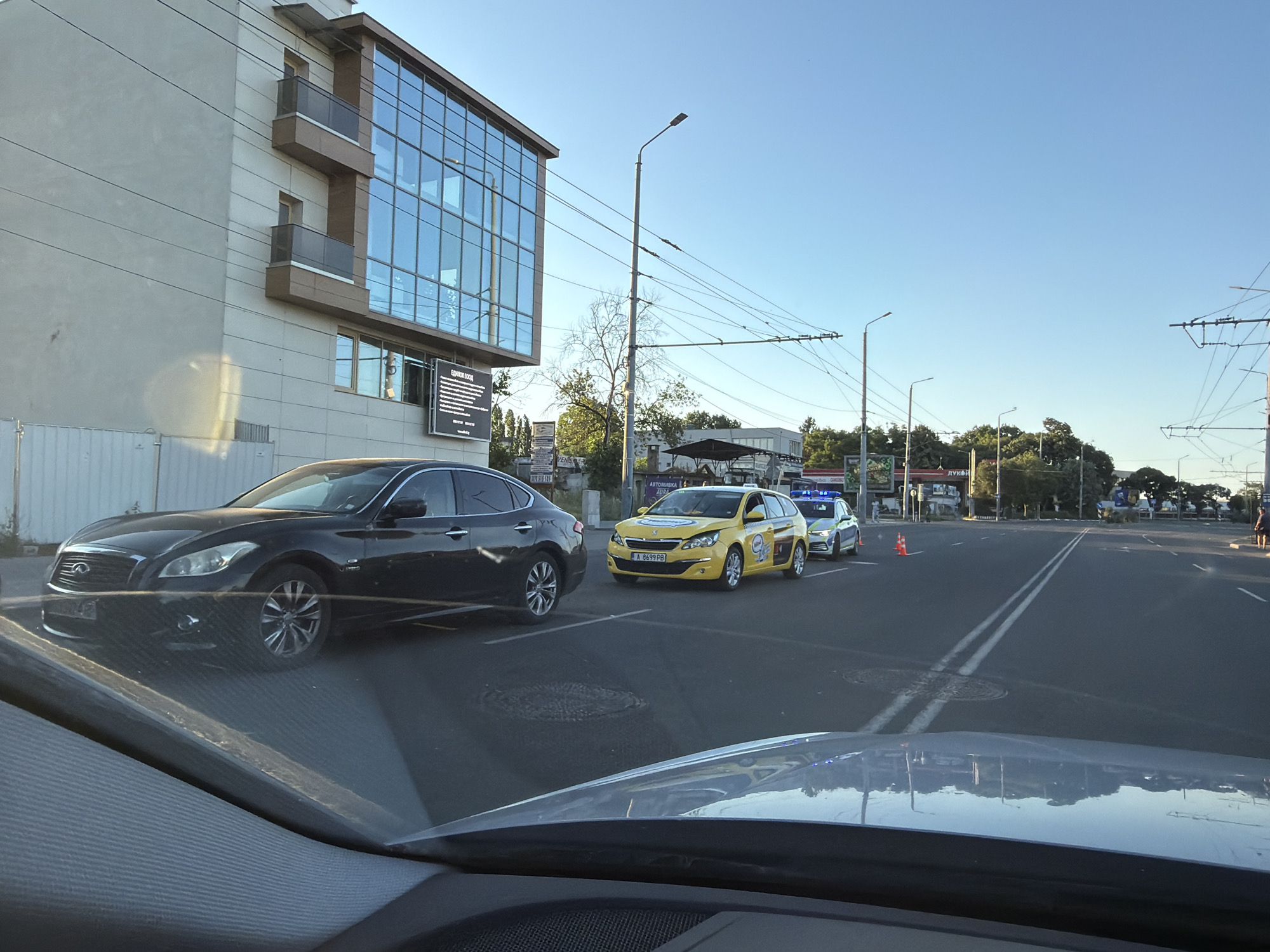 The image depicts a street scene in Burgas, Bulgaria, under a clear blue sky. In the foreground, the hood and windshield of a car are visible, reflecting the surrounding environment. A black Infiniti sedan is parked on the left side of the road. Further down the street, a yellow taxi, identifiable by its markings, is stopped, followed by a police car. Orange traffic cones are placed on the road, suggesting a temporary obstruction or a controlled area. Several streetlights and overhead wires for public transportation are present. A modern building with a glass facade stands on the left, with balconies protruding from its upper floors. Trees and other structures line the background, indicating an urban setting. The overall impression is a snapshot of a normal day in the city, with a slight indication of a traffic management situation.
