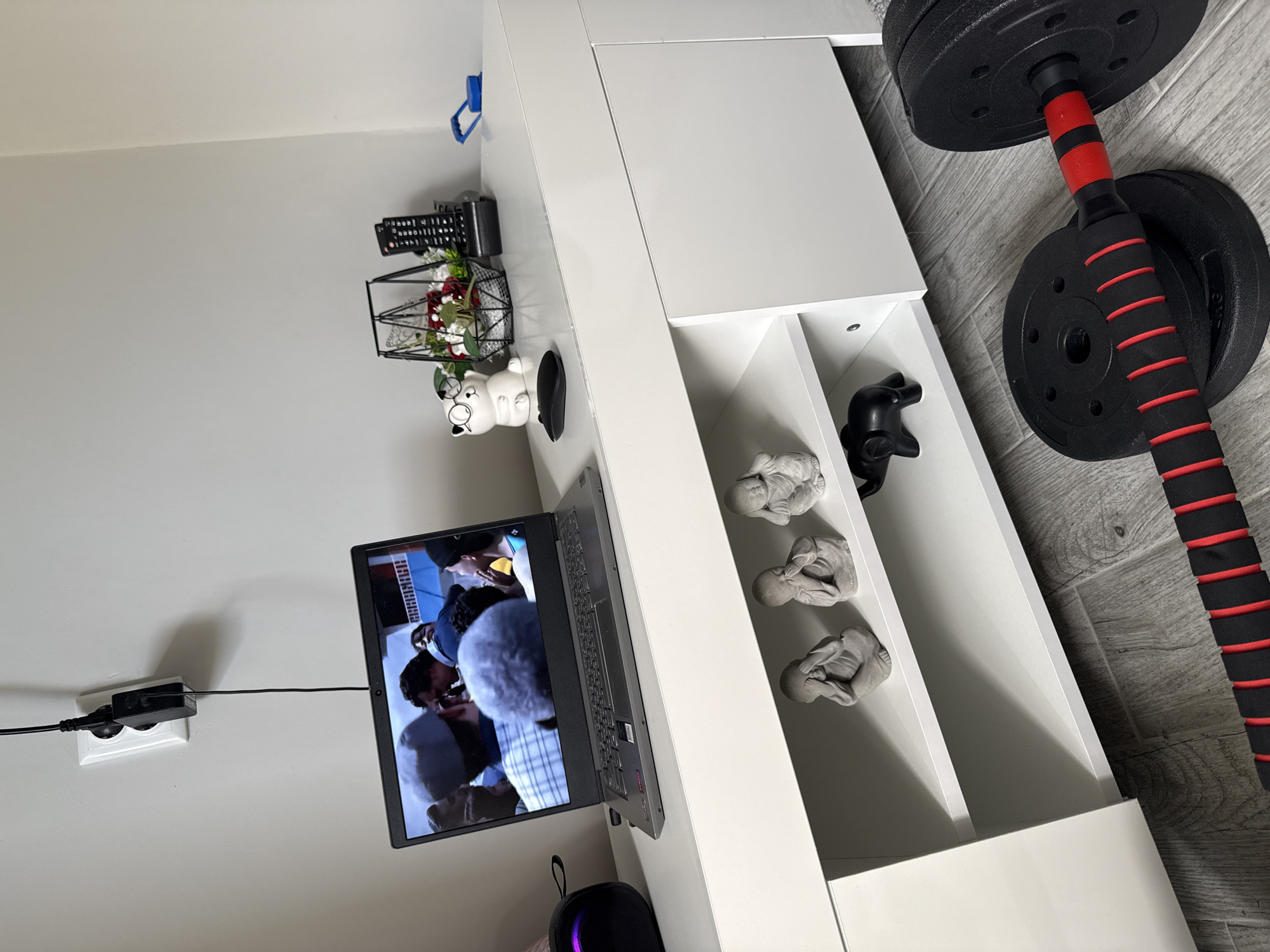 The image presents an interior setting. A laptop displaying a video is positioned on a white cabinet or shelf. The cabinet has open compartments, one containing three small, gray, abstract statues of meditating figures and another compartment with a black game controller. On the cabinet's surface, there's a collection of items including a remote control, a small wire basket with flowers, a piggy bank wearing glasses, and a computer mouse. A black and red dumbbell rests on the floor beside the cabinet. The scene is domestic and likely indoors.
There is no information in the image that indicates that the location is Stara Zagora, Bulgaria.