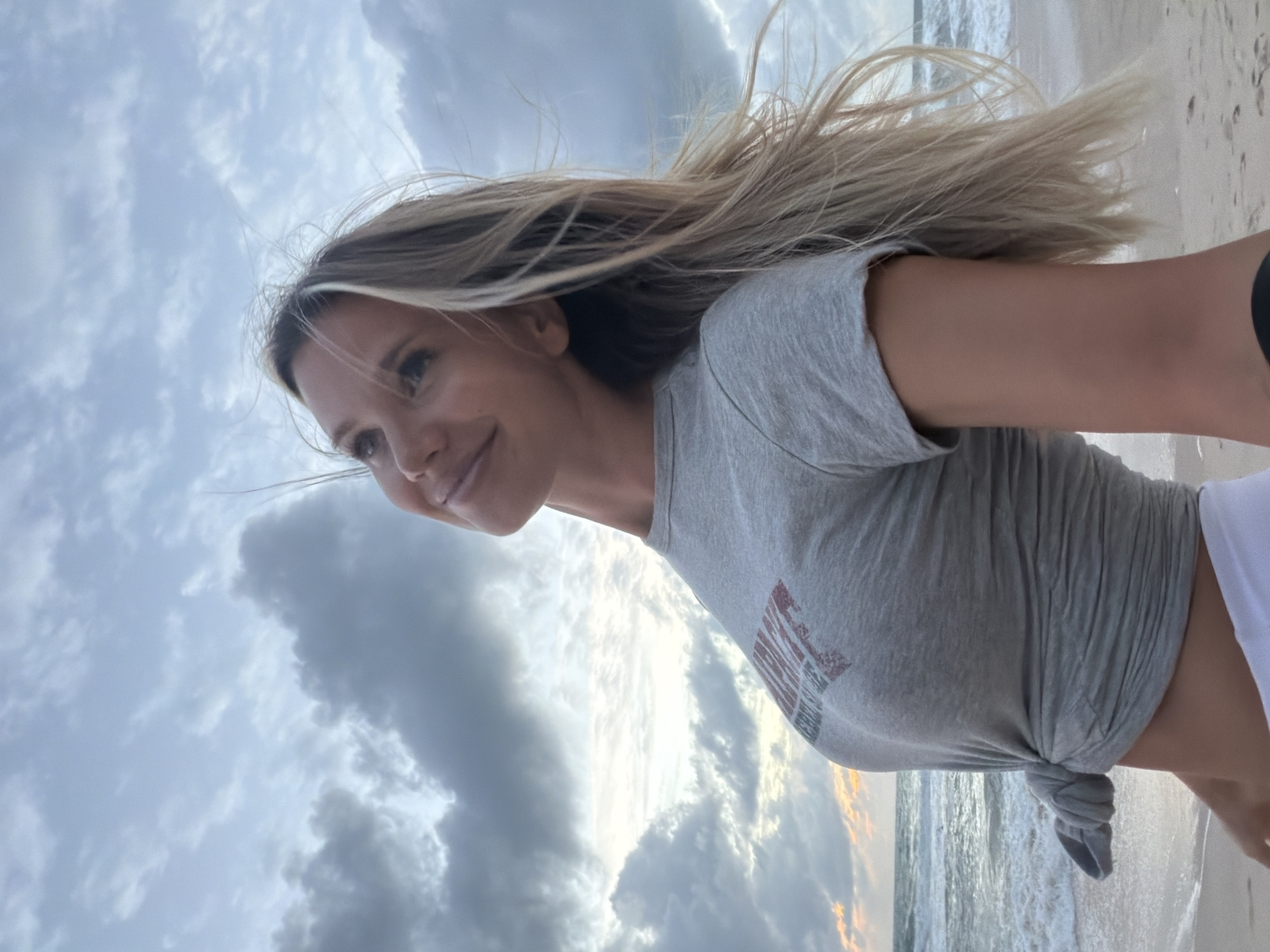 A medium shot captures a blonde woman on a beach, looking up and to her left with a slight smile. She is wearing a grey t-shirt knotted at the waist, exposing her midriff, and white shorts. Her long blonde hair is windswept. In the background, the ocean with moderate waves meets a sandy beach. The sky is cloudy with hints of sunrise or sunset. The location is identified as Choeng Thale, Thailand.