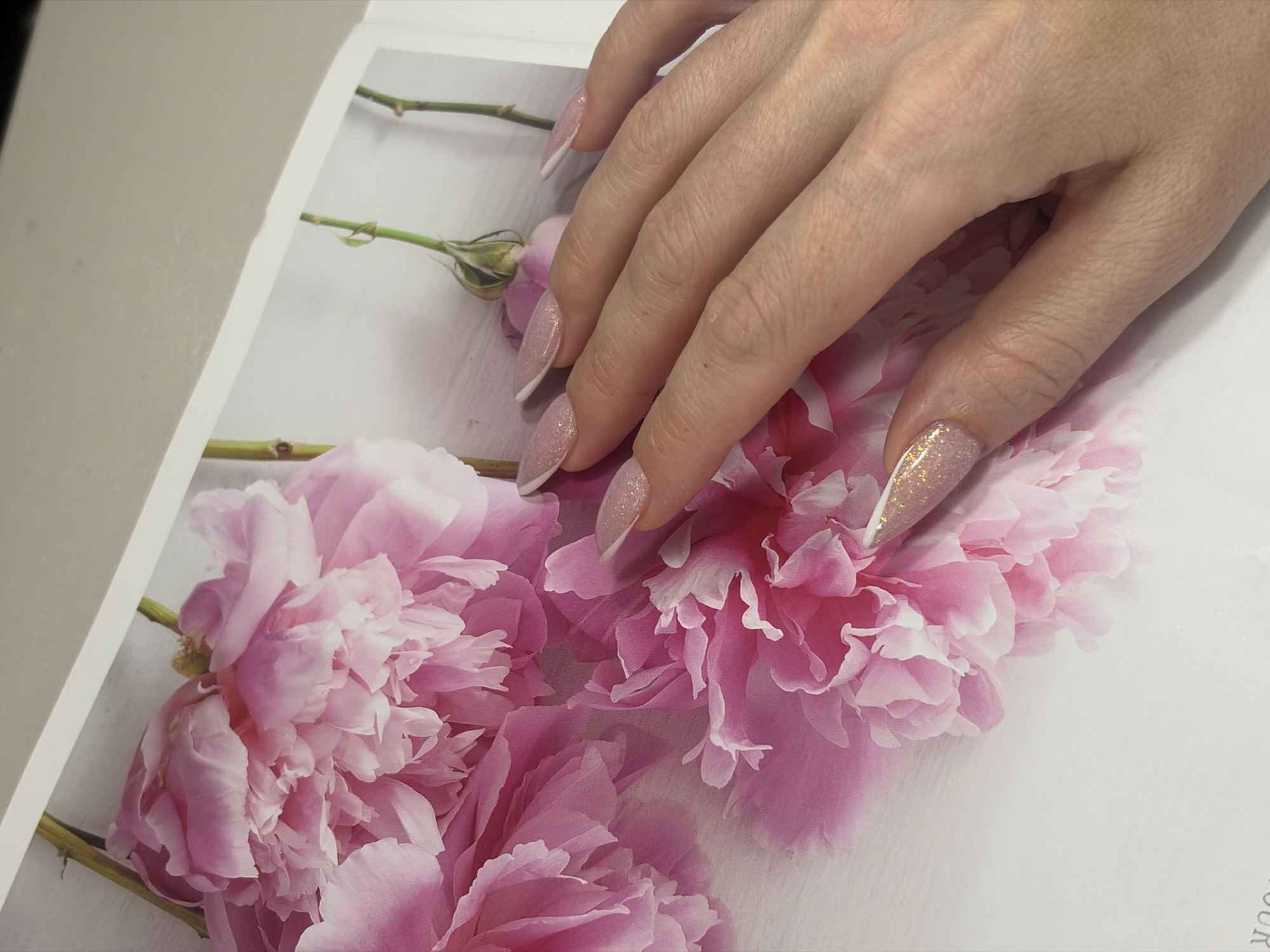A close-up photograph shows a woman's hand with manicured fingernails resting on a surface printed with pink peony flowers. The nails are long and pointed, with a light pink base and white tips, some adorned with glitter. The background is a light surface with pink peony flower prints. The image does not provide sufficient information to determine a specific location within Saint Petersburg, Russia.