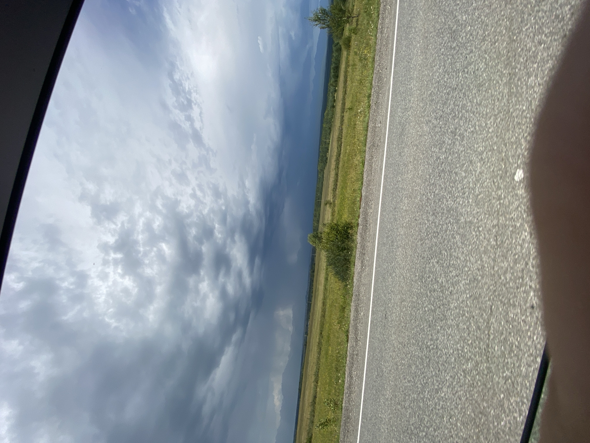 The image shows a view from a vehicle traveling on a paved road with a white line marking the lane. The road is in the foreground, with a wide, green, grassy field extending beyond it. In the distance, there are trees and mountains under a dramatic, cloudy sky, with dark storm clouds in the center and lighter clouds on the sides. The location is the city of Zaktui, Russia. There are no people or animals visible in the image.
