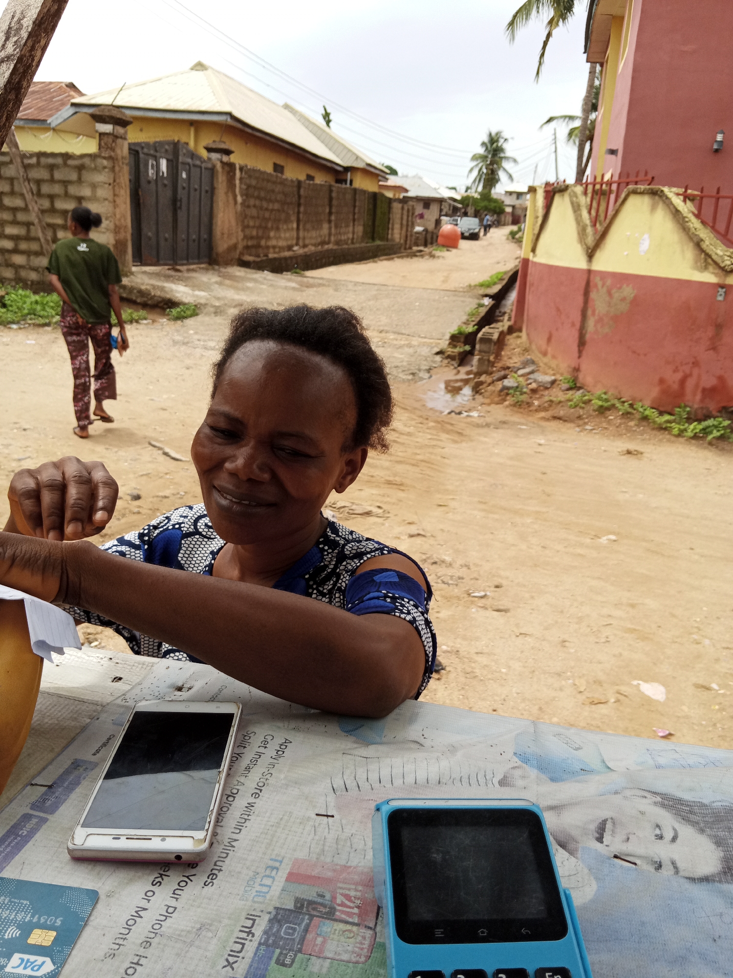 The image presents a medium shot of an African woman, likely in her 50s, smiling and interacting with something off-camera to the left. She is wearing a blue and white patterned top and has dark hair styled close to her head. In front of her, on a table covered with newspaper advertisements for mobile phones (TECNO and Infinix), are a white smartphone, a blue Point of Sale (POS) terminal, and what appears to be a PAC bank card.
In the background, the scene extends into a residential street, with buildings on either side painted in yellows and reds. A person wearing a green shirt and patterned pants is walking away from the camera on the left side of the road. Further down the street, a vehicle is visible. The road itself is unpaved. The setting appears to be a street scene in an urban or peri-urban area.