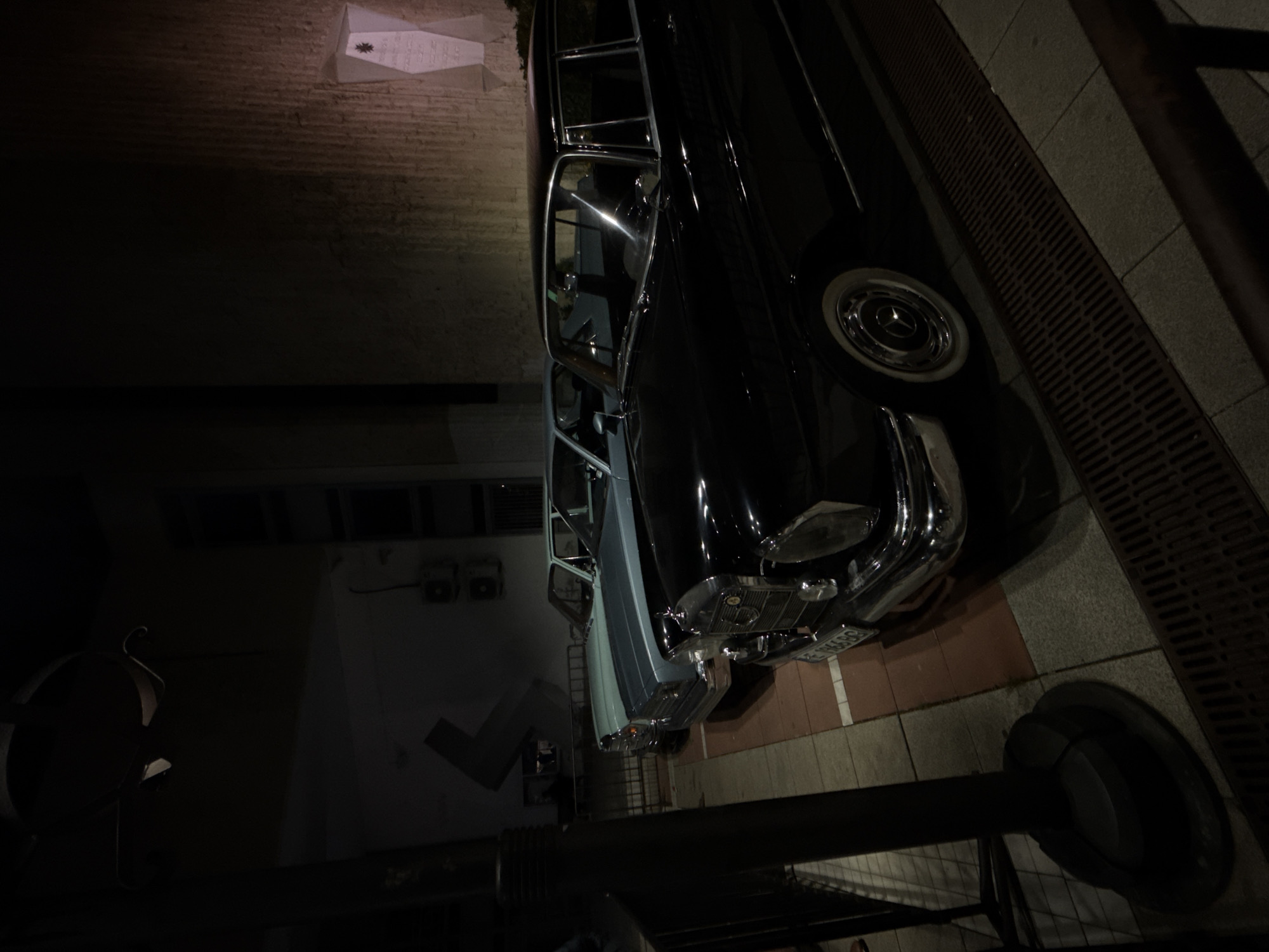 A black Mercedes-Benz car is parked on a tiled sidewalk next to a building. The location is Blagoevgrad, Bulgaria. The car is parked near a black metal pole and fence. The environment appears to be a street at night.