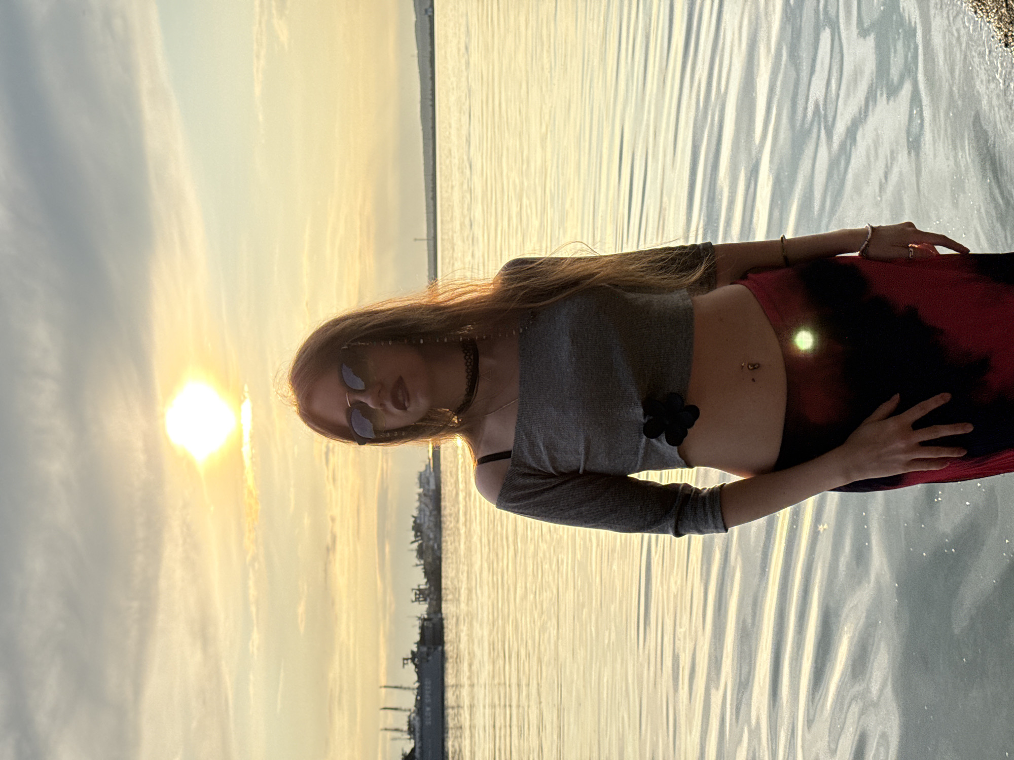 A young woman stands by the water with ships docked in the distance. The sun is low in the sky, casting a warm glow on the scene. She is wearing a gray crop top, sunglasses, and a long skirt. Her hair is long and brown, and it falls around her shoulders. The water is calm and reflects the colors of the sky. The overall atmosphere is peaceful and serene.
