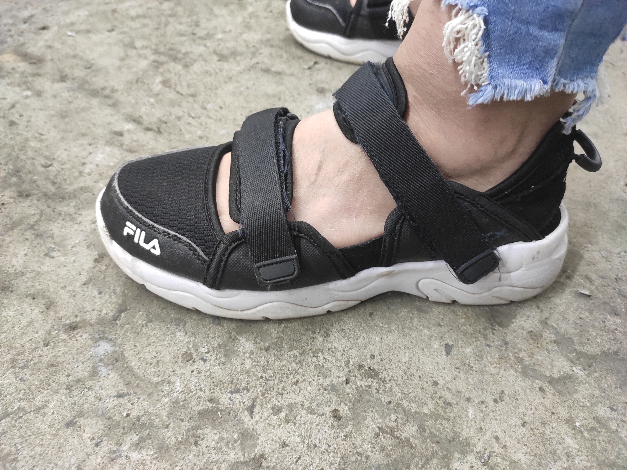 A black Fila sandal with white trim is worn on a person's foot, which is resting on a concrete surface. The sandal has two adjustable straps across the top of the foot. Frayed denim jeans are visible above the sandal. The concrete surface appears worn and has a textured appearance, with some scattered debris. The scene is outdoors, likely on a sidewalk or paved area. The location is not specified in the image or metadata.