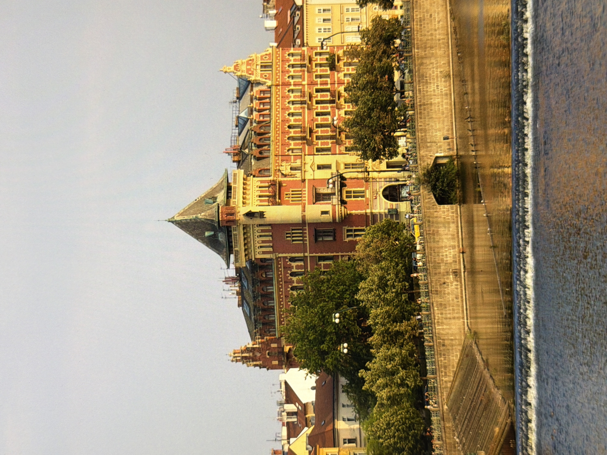 This is a daytime, eye-level, medium shot of the embankment and a building on the right side of the Vltava River in Prague, Czech Republic. The building is multi-story with a red facade and yellow trim, featuring a steeply pitched roof with dormer windows. There are trees in the foreground along the embankment, and a stone wall separates the walkway from the river. The river is calm with a small waterfall or dam in the foreground. People are visible walking along the embankment and some vehicles are present. The sky is clear and bright.
