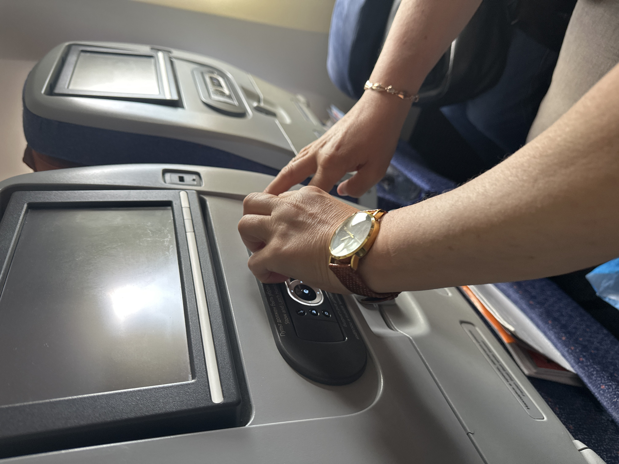The image shows a close-up view of an airplane cabin. In the foreground, a person's arm wearing a gold-faced watch with a brown leather strap is visible. Their hand is touching a control panel on the back of an airplane seat. The control panel has a screen and several buttons. Text in Cyrillic and English on the panel reads "Пульт должен быть убран" and "Stow and Latch Handle". Another hand, wearing a delicate gold bracelet, is also visible reaching towards the control panel. The background shows the backs of airplane seats, with one seat featuring a small screen. The environment appears to be a typical airplane interior, with blue upholstery and grey plastic elements. There is no indication of the city of Nochvino, Russia, within the image.