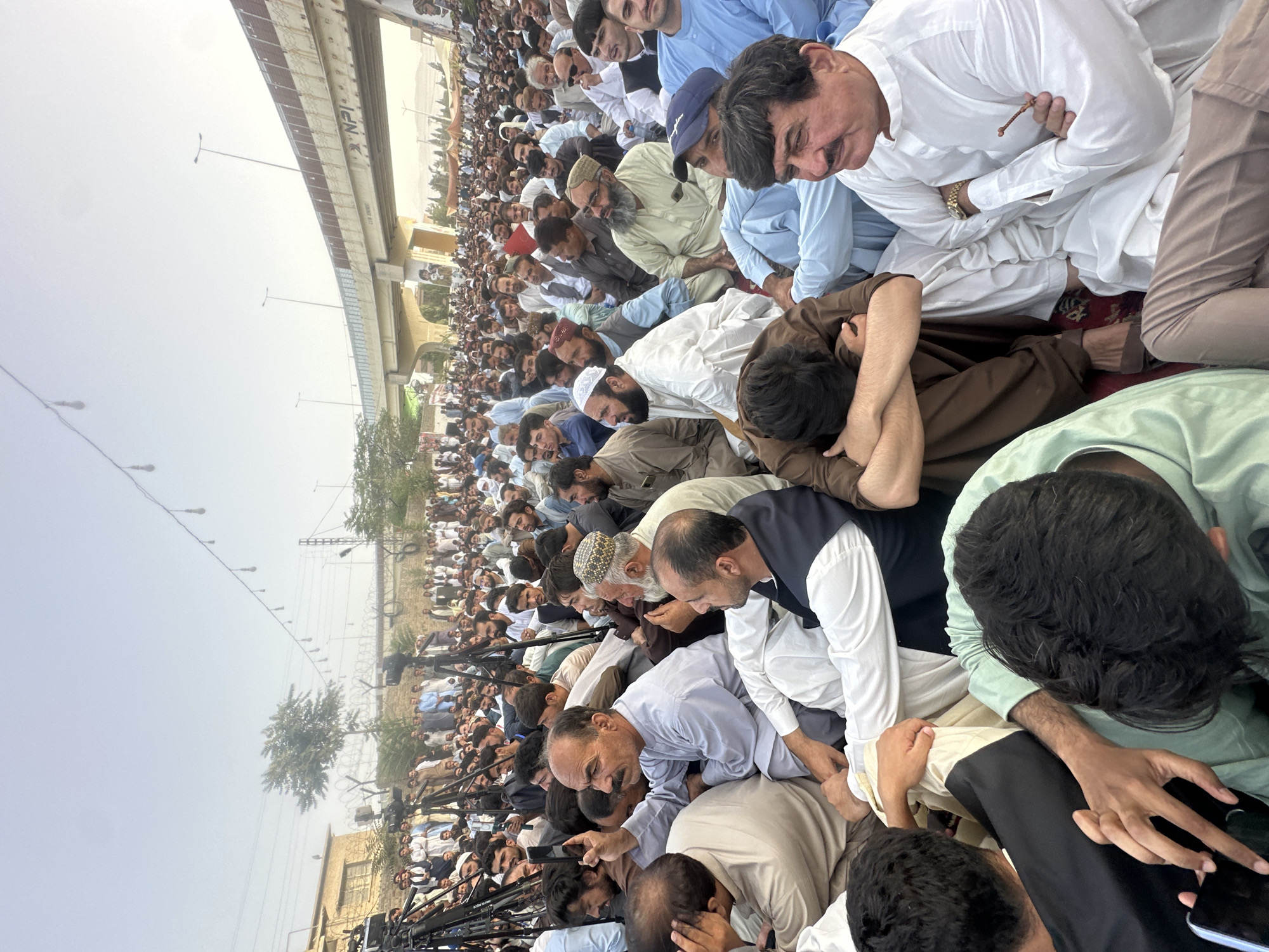 A large crowd of men are gathered outdoors in Quetta City Tehsil, Pakistan, seated on the ground. Many are dressed in traditional clothing, with some wearing white shalwar kameez and others in darker colors. Several men are looking at their phones, and some are holding them up, suggesting they are recording or photographing the event.
In the foreground, a man in a light blue shalwar kameez with a white vest is looking to his right and holding prayer beads. To his left, another man in a white shalwar kameez has his arms crossed and is looking forward. In the middle ground, a man in a light gray shalwar kameez is looking up and holding his phone.
The background reveals a street scene with a bridge overhead. Several cameras on tripods are visible among the crowd, indicating media presence. The sky is overcast, and the overall lighting suggests it is daytime. The environment appears to be an urban setting, possibly a public gathering or demonstration.