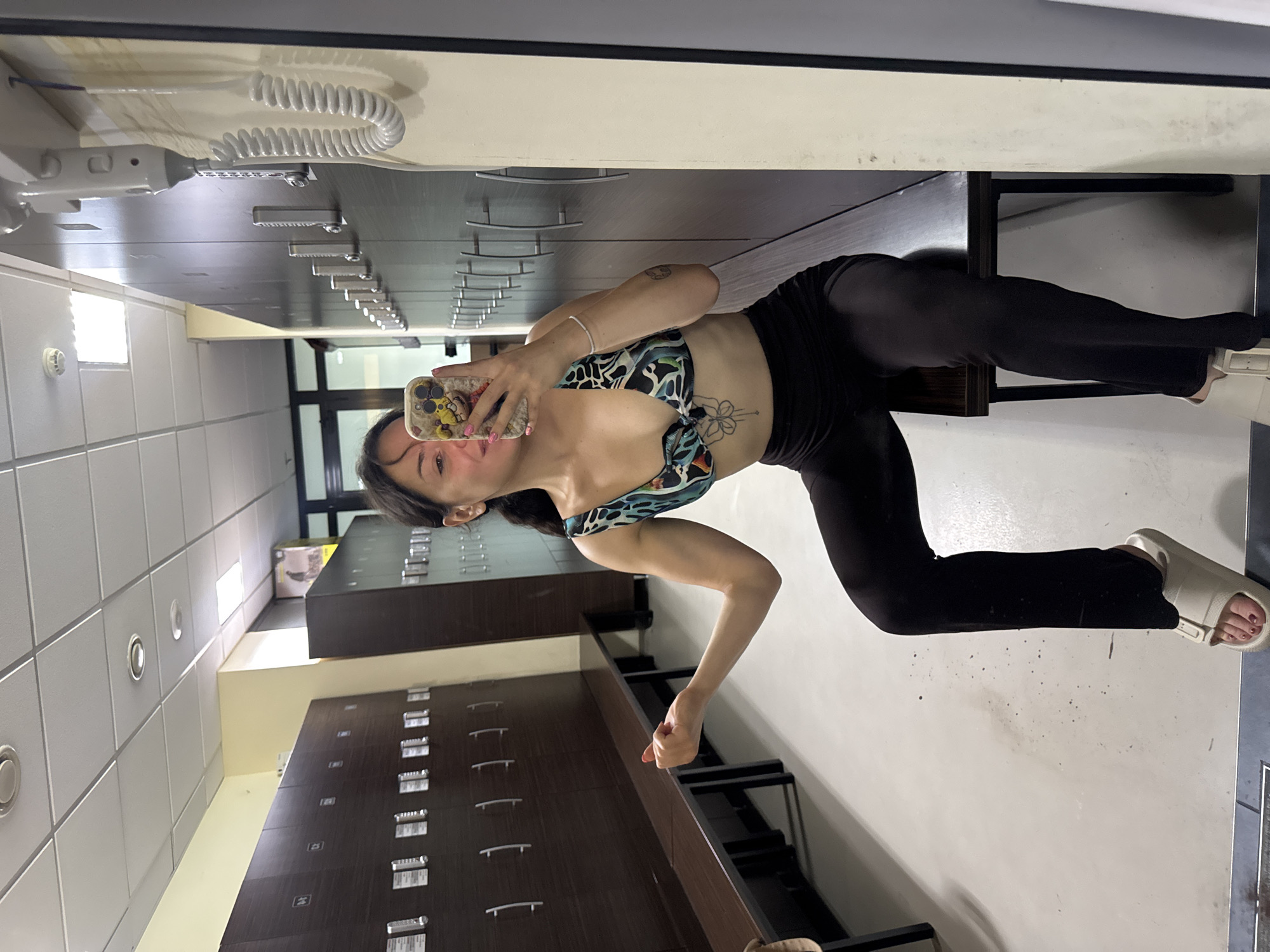 A woman is taking a mirror selfie in a room that appears to be an office or storage area. She is wearing a colorful patterned sports bra and black leggings. Her pose is dynamic, with her right leg extended back and her left leg bent. She is holding a smartphone in her right hand, capturing the reflection.
The background shows rows of filing cabinets, suggesting an administrative or archival setting. The lighting appears to be artificial, indicating an indoor environment. There is a coiled corded object on a shelf in the upper left corner, possibly a telephone or medical equipment. The overall impression is of a casual, private moment captured within a functional space. The location context is Burgas, Bulgaria, but there are no specific visual cues within the image to confirm this.