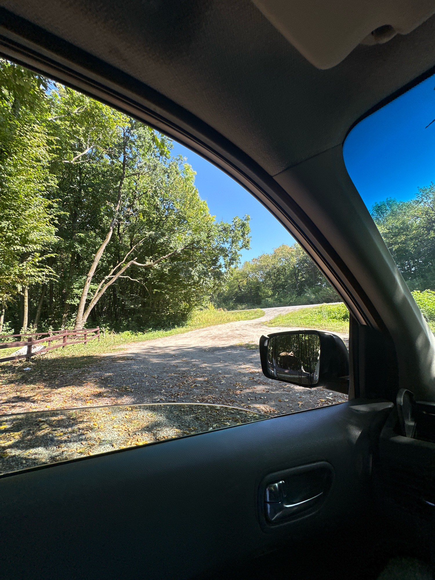 The image, captured from inside a vehicle, shows a dirt road curving through a wooded area under a clear blue sky. Sunlight dapples the road, which is covered in fallen leaves. A brown wooden fence runs along the left side of the road, bordering a dense forest of green trees. In the rearview mirror, a reflection shows the trees and sky. The interior of the car, including the door panel and window frame, is visible in the foreground. The scene appears to be located in a rural or natural setting, likely on the outskirts of Shatava, Ukraine.