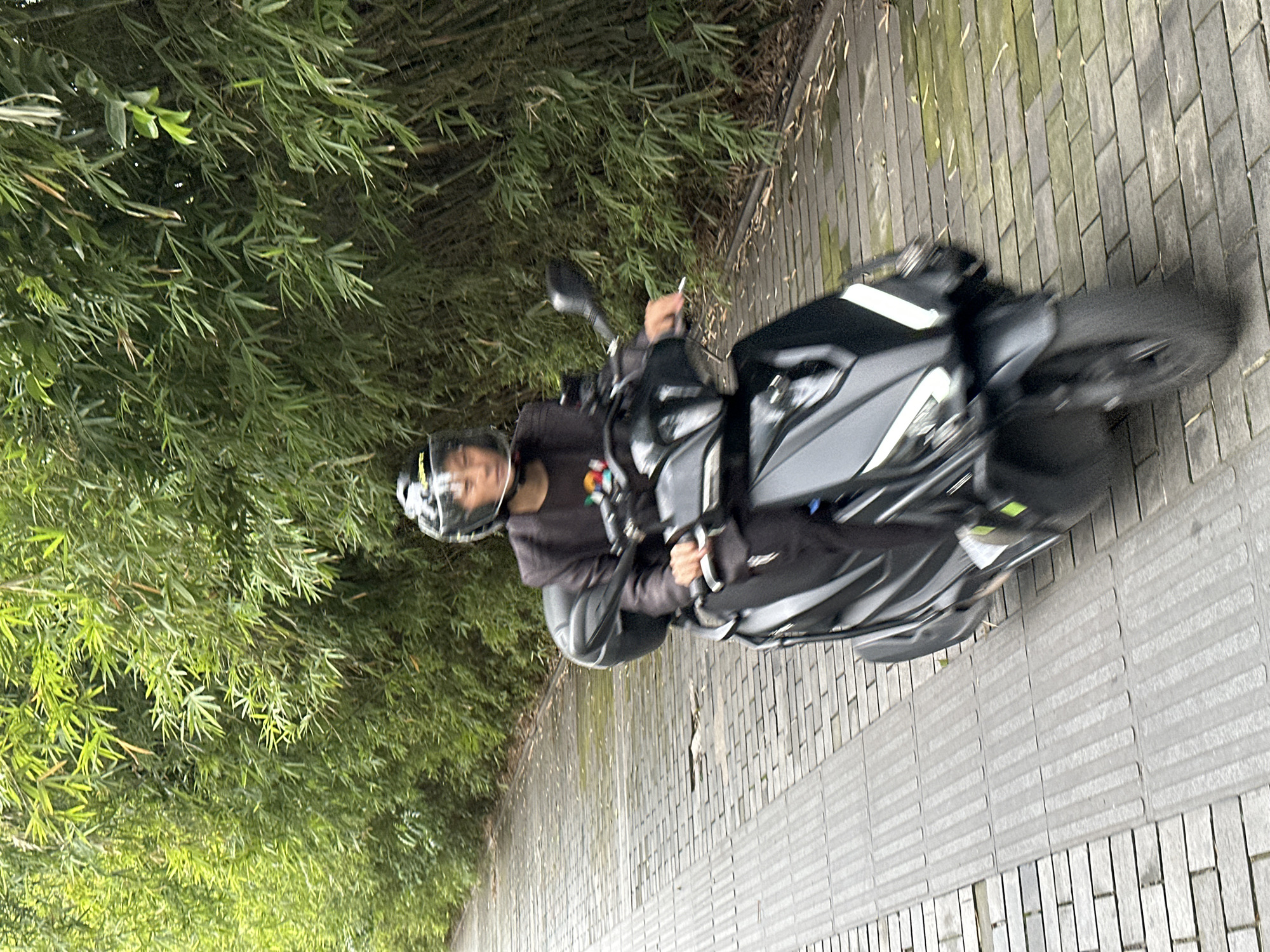 A person wearing a helmet is riding a black scooter on a paved walkway lined with bamboo. The walkway appears to be wet. The photo was taken in Liangjiang, China.