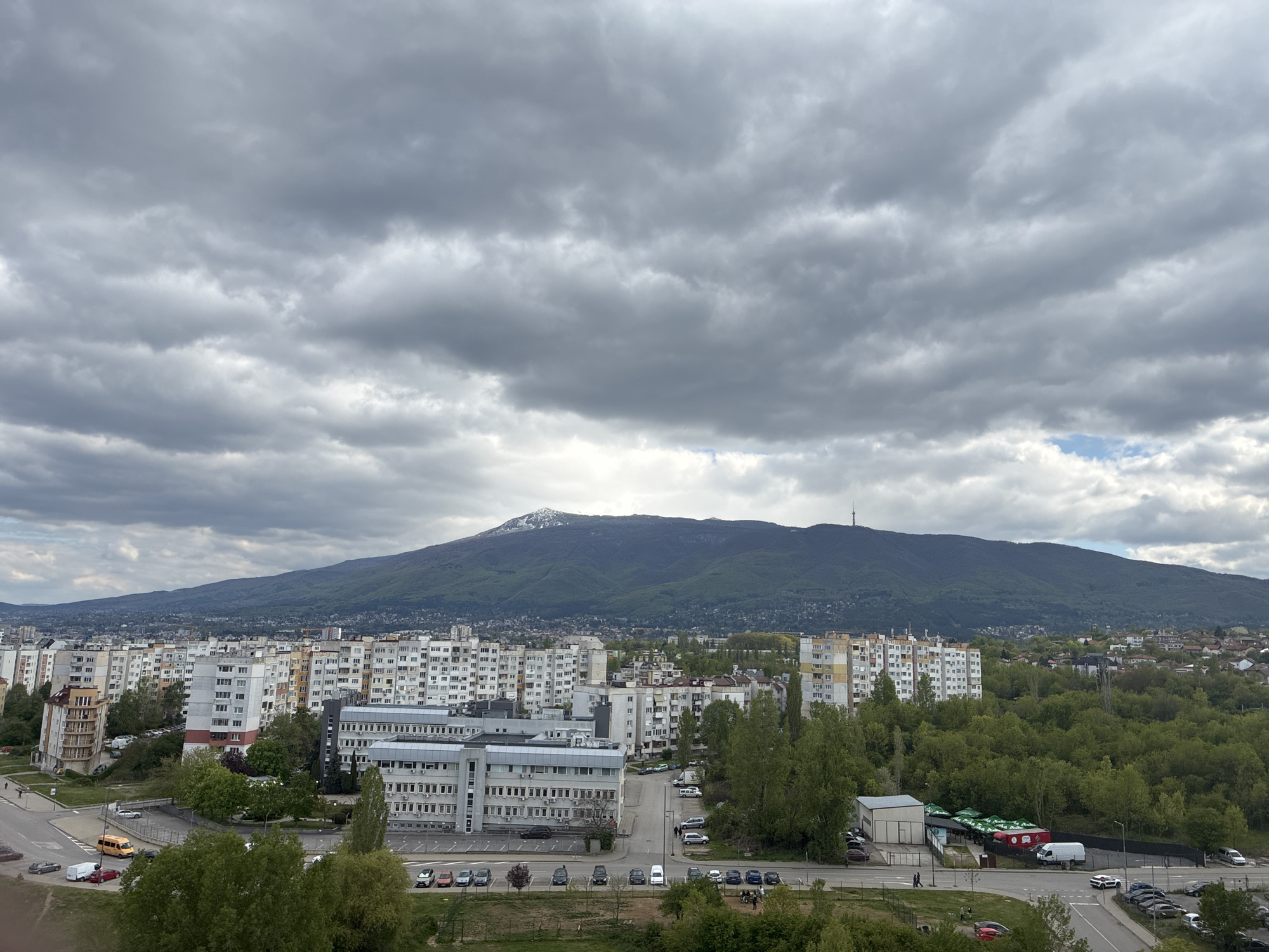 The image captures a cityscape in Sofia, Bulgaria, set against the backdrop of Vitosha Mountain, which has a snow-dusted peak and a prominent communication tower near its summit. The sky is heavily overcast with dark, gray clouds. In the foreground, there are several apartment buildings of varying heights and architectural styles, along with office buildings featuring rows of windows. Roads intersect, with parked cars visible along the streets and in parking areas. Trees with green foliage are interspersed among the buildings, adding some color to the otherwise muted palette of the scene. A few people can be seen walking along the sidewalks. The overall impression is one of an urban environment on a cloudy day.