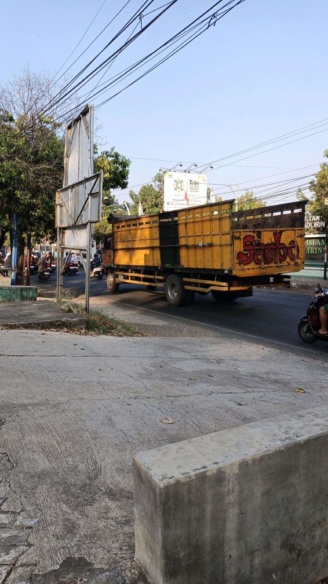In the city of Kuwasen, Indonesia, a bright yellow truck with black accents is captured driving on a paved road under a clear blue sky. The truck has a large, open cargo bed with wooden planks on the sides and a metal frame at the top. Red graffiti reading "Sambel" is visible on the rear right side of the truck. Several motorcycles are visible on the left side of the road, some with riders, suggesting a traffic scene. A large, white billboard stands behind the truck, partially obscured by trees. The road is lined with trees on the left, and there is a concrete sidewalk and curb in the foreground. The overall atmosphere suggests a typical day in an Indonesian urban or semi-urban environment.