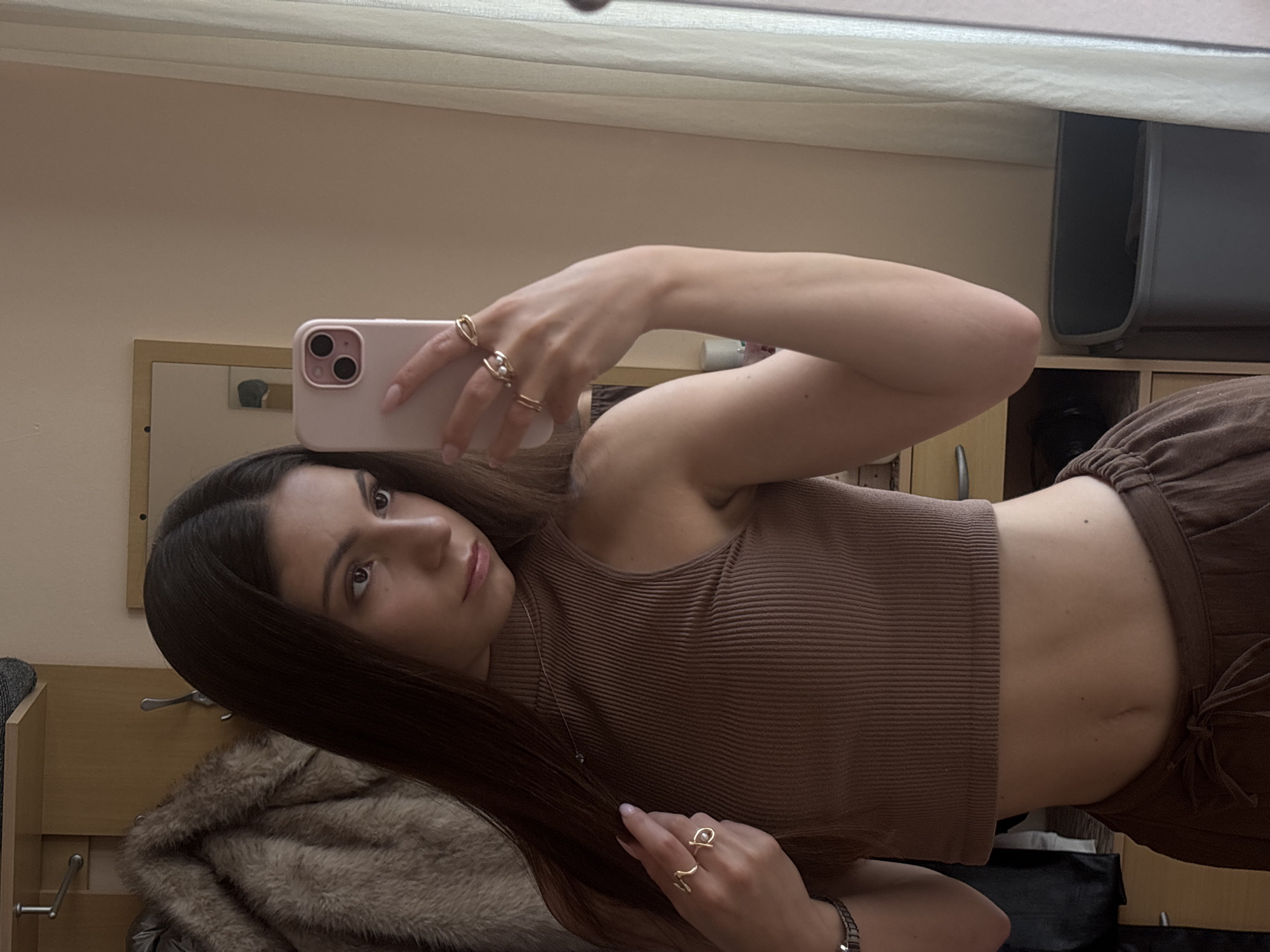 The image is a selfie taken by a young woman indoors, with the camera angled upwards. She has long, dark brown hair and is wearing a brown, ribbed, sleeveless crop top and brown drawstring pants. She is also wearing several rings on her fingers. The background shows a mirror, a closet with a fur coat on a hanger, and part of a room with wooden furniture. The lighting suggests it is daytime, but the specific time is not discernible. The overall mood is casual and relaxed. No visible text is present in the image.