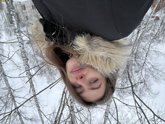 This image depicts a female individual outdoors in Chaikovsky, Russia, during winter conditions. The subject, a young adult with long brown hair, is positioned in the foreground, captured in a low-angle close-up, looking slightly upwards and towards the viewer. She is wearing a black winter parka with a prominent light-colored, likely faux fur, trim around the hood, which, along with her hair, is visibly dusted with fine snowflakes. Her right eye, nose, and the right side of her mouth are clearly visible, with her lips showing a slight upward curve. The mid-ground is characterized by multiple bare birch trees, their white bark with dark markings and branches lightly covered in snow. The sky is uniformly overcast and light grey. In the background, partially obscured by the trees, multi-story residential buildings are visible, also covered in snow, indicating an urban or suburban setting within Chaikovsky.