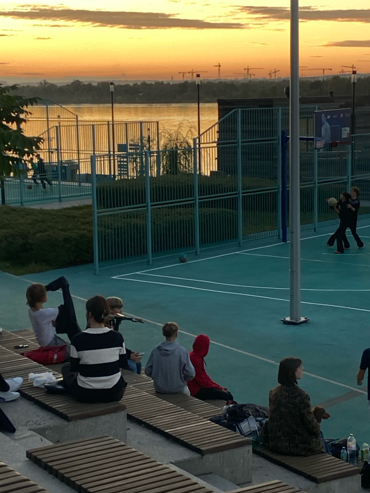 The image depicts an outdoor basketball court at dusk, with a body of water and distant construction cranes in the background. The sky is illuminated by a warm sunset. Several individuals are present: two people are playing basketball on the court, one with a ball in hand. On the bleachers overlooking the court, a group of people are seated, some observing the game and others engaged in their own activities. One person on the bleachers is holding a small dog. The setting appears to be a public recreational area, possibly in or near the city of Perm, Russia, given the context of distant construction and the overall urban landscape. The lighting suggests it is early evening, with the sun setting on the horizon.