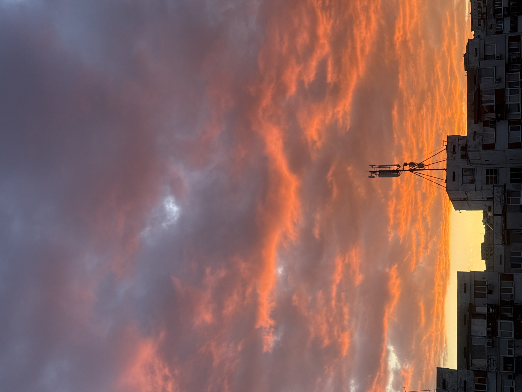 The image shows a sunset over a city. The sky is filled with orange and red clouds. On the right side of the image are tall buildings, likely apartment blocks, with numerous windows and some air conditioning units visible. In the foreground is a telecommunications tower mounted on top of one of the buildings. Based on the architectural style of the buildings, this image is most likely taken in Sofia, Bulgaria.