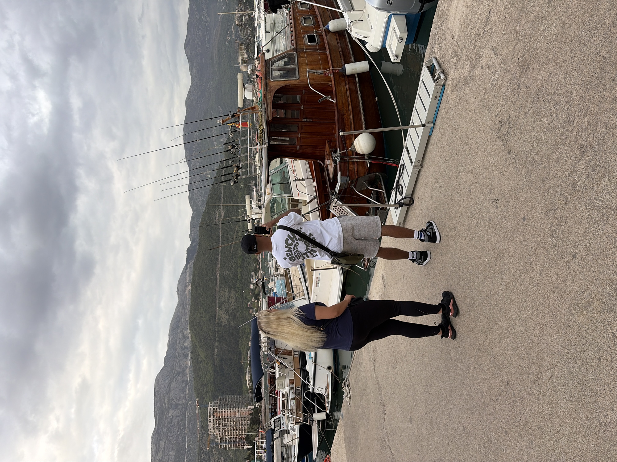 The image shows two individuals, a man and a woman, standing on a concrete pier in Budva, Montenegro, alongside several boats docked in the harbor. The man is wearing a white T-shirt, gray shorts, black and white sneakers, and a black baseball cap. He has a small bag slung across his shoulder and appears to be looking at something off-camera. The woman is wearing a navy blue T-shirt, black leggings, and black sneakers. She has long blonde hair and is also looking towards the boats.
Behind them, a wooden boat with numerous fishing rods attached to the back is visible. There are other boats docked in the harbor, with parts of buildings visible in the distance. A large mountain is in the background, under a cloudy sky. The ground is concrete with visible wear and tear.