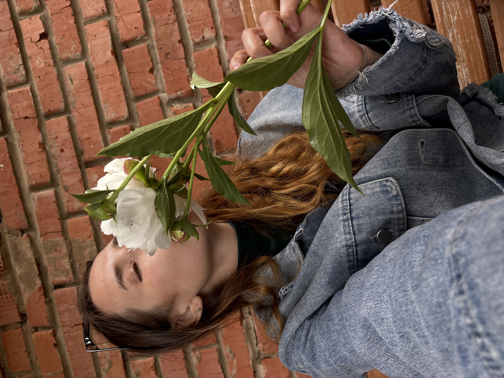 A young woman with long, wavy brown hair is pictured from a high angle, sitting on a wooden bench in front of a brick wall. She is wearing a denim jacket over a dark green turtleneck. Her right hand holds a white peony flower, bringing it to her nose to smell. She is wearing glasses on top of her head. The brick wall shows signs of age, with some cracks and uneven mortar. The image was taken outdoors, possibly in a park or courtyard in Votkinsk, Russia.