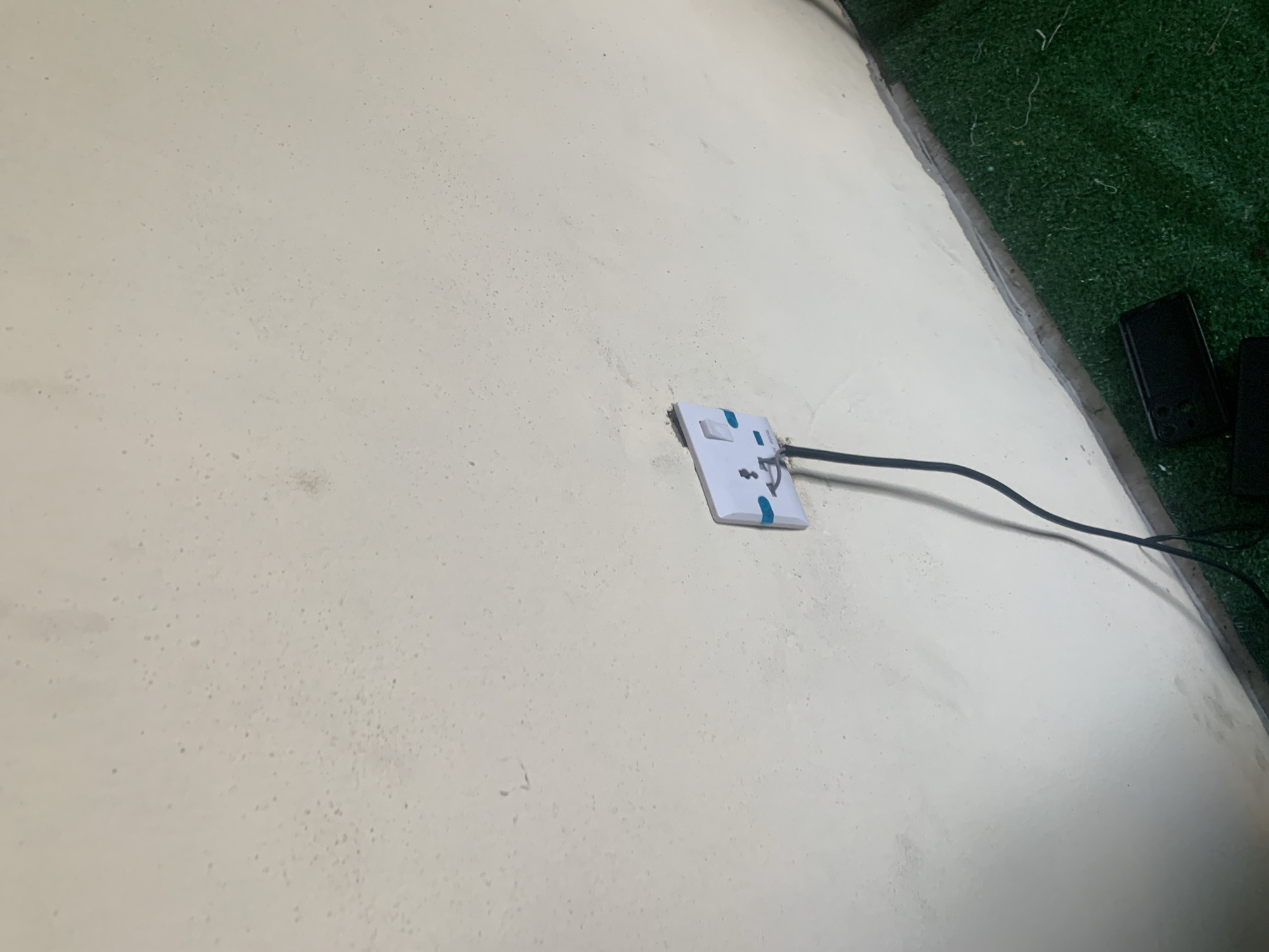 In Jauro Yinu, Nigeria, an electrical socket is mounted on a textured, cream-colored wall. A black electrical cord extends downwards from the socket. The wall is slightly discolored with small, dark spots. A narrow strip of grey material runs along the bottom of the wall. Below this, a patch of vibrant green artificial turf is visible. On the turf, near the wall, a black rectangular object, possibly a phone, lies flat. The lighting in the scene casts a shadow from the electrical cord onto the wall.