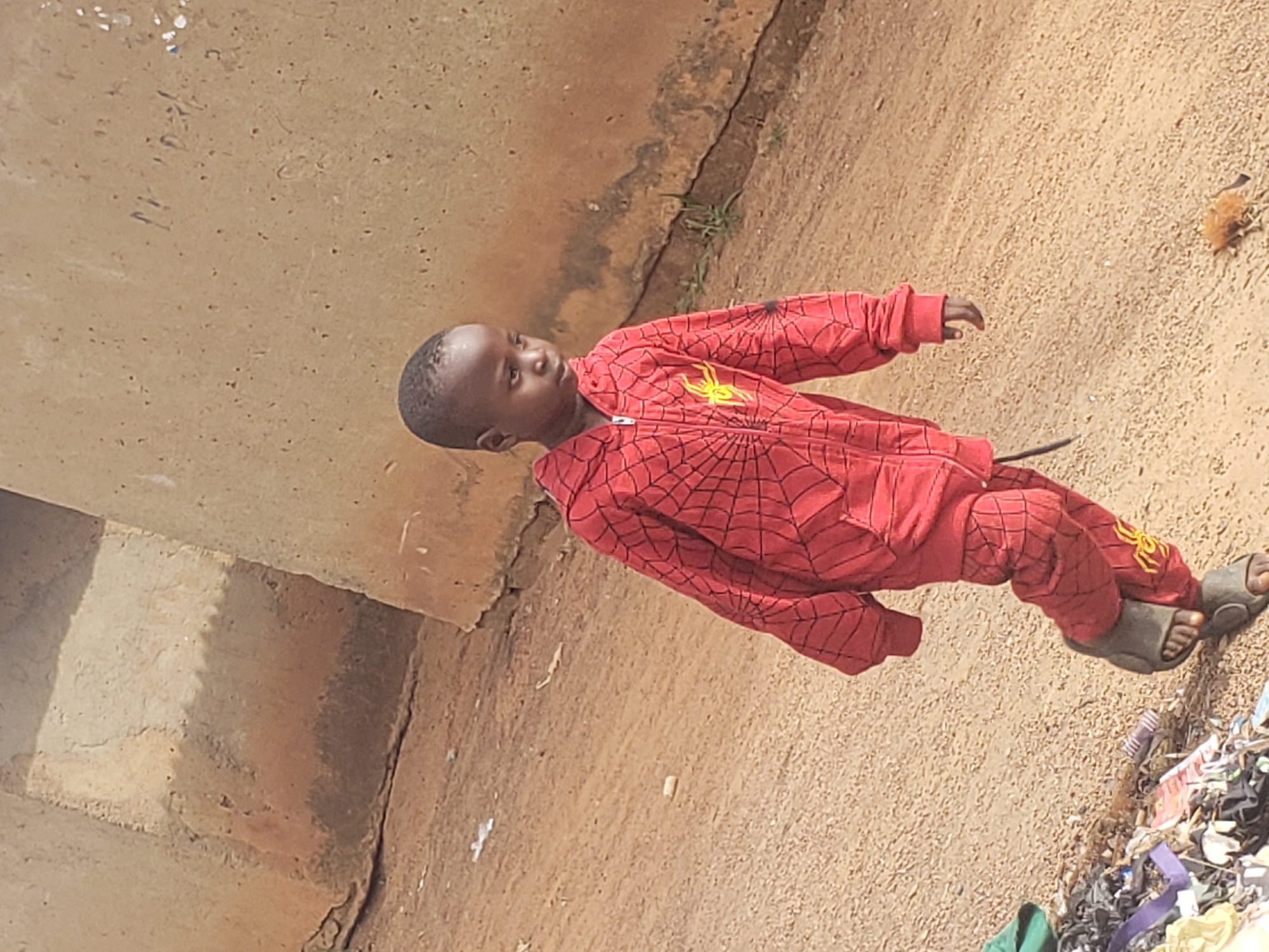 In Kachia, Nigeria, a young boy with dark skin and short, dark hair walks on a dirt path. He is wearing a red hooded tracksuit with a black spiderweb pattern and a yellow spider emblem on the chest and legs. The boy is looking to his right, with his left arm bent at the elbow and his right hand by his side. His feet are shod in dark sandals. To his left, there is a pile of assorted trash. In the background, a rough, textured wall made of earth and stone rises, with a prominent crack running horizontally across it. The ground is composed of loose dirt and small pebbles.