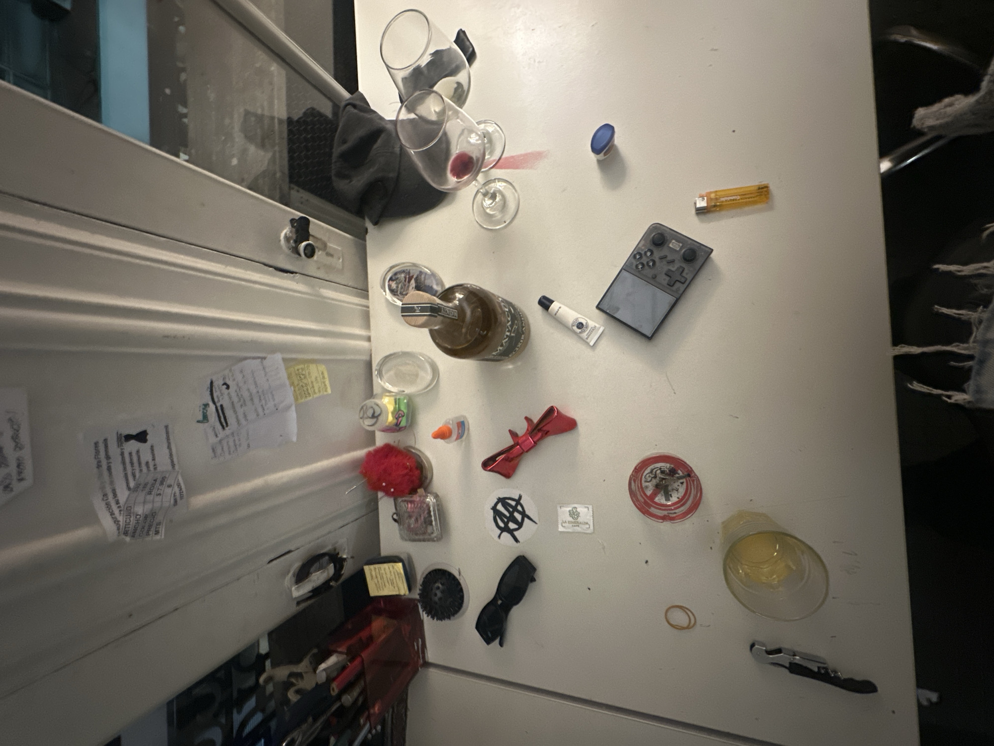 The image displays a cluttered white surface, likely the top of a refrigerator or a counter, seen from an angled overhead perspective. To the left, a partially visible white appliance door is adorned with several papers held by magnets.
On the main white surface, in the upper left, two clear wine glasses are present; one stands upright, and another is inverted, resting next to a distinct red wine stain. A third inverted wine glass with a similar stain is positioned slightly below. A black fabric item, possibly a cap or cloth, is also in this area.
Towards the top right, a small blue-capped container, a yellow disposable lighter, and a grey and black handheld controller-like device are visible. In the central area, a brown glass bottle with a wooden stopper, labeled "MATERIA ELIXIR," lies horizontally. Adjacent to it is a small white tube with a blue cap, bearing the text "L'OCCITANE."
On the vertical surface to the left, which appears to be a refrigerator door, various items are attached. These include a red fuzzy pom-pom, a small bottle of glue, and a small clear container. A red metallic bow, black sunglasses, and a circular black object are also present. Two stickers are visible: one depicting an anarchy symbol (an 'A' within an 'O'), and another rectangular sticker for "LA ESCONDIDA Cafe." Several papers, possibly receipts or notes written in Spanish, are held by magnets; fragments of text like "ARTICULO," "PRECIO," "ROSA," and "MTR" are discernible.
On the main white surface towards the bottom right, a clear glass contains a small amount of yellow liquid. A round ashtray featuring a "no smoking" symbol, a rubber band, and a silver and black corkscrew complete the arrangement of objects.
The scene captures a collection of everyday items in a domestic setting, suggesting recent casual use. No specific activity is taking place, and no individuals are present. The scattered nature of the objects gives an impression of casual disarray. Given the Buenos Aires, Argentina location context, the Spanish text on the papers is consistent.